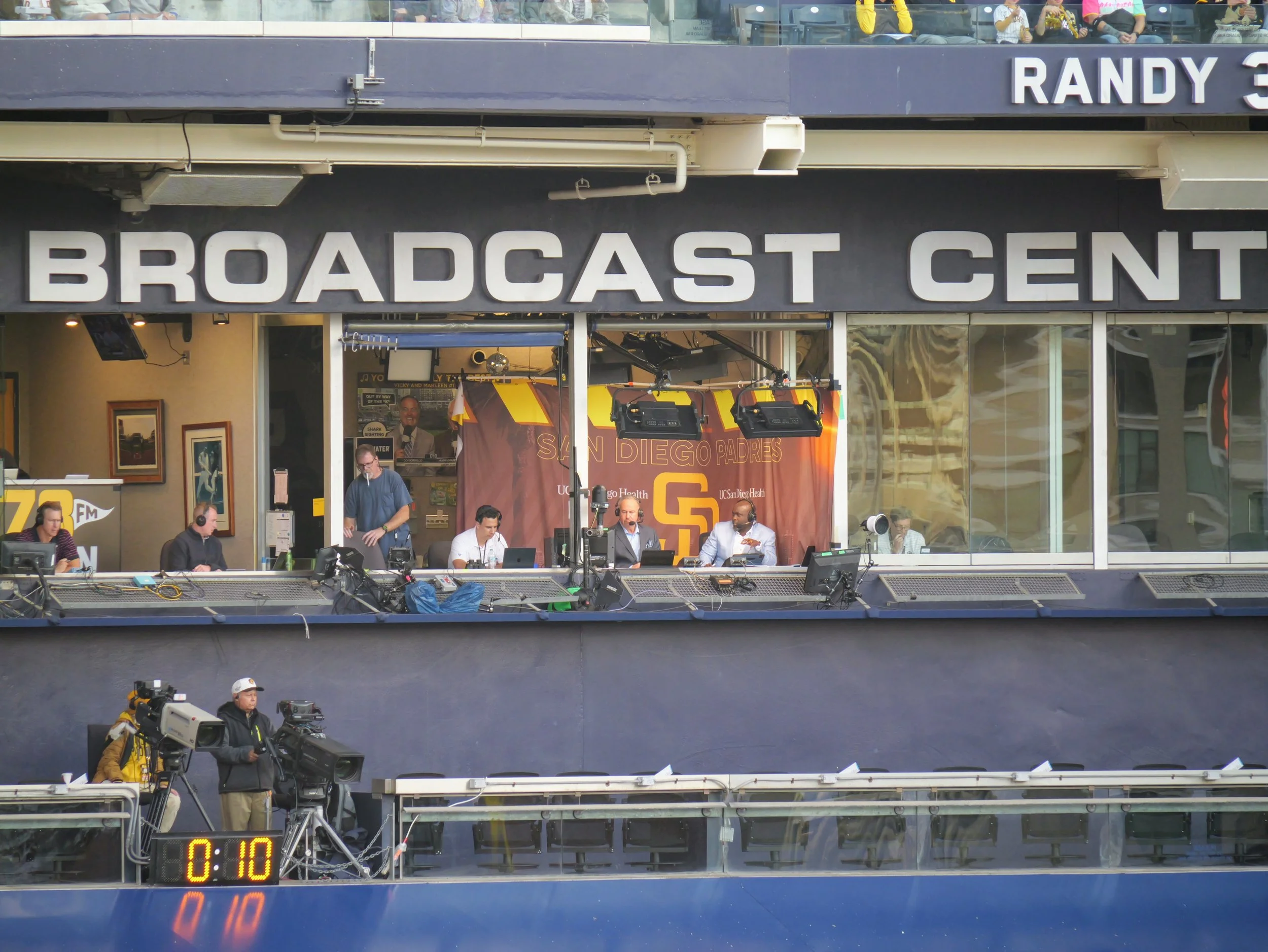 A broadcast booth at a sporting event with people working at computers, a camera operator, and an audience in the background. The sign reads 'Broadcast Center'.