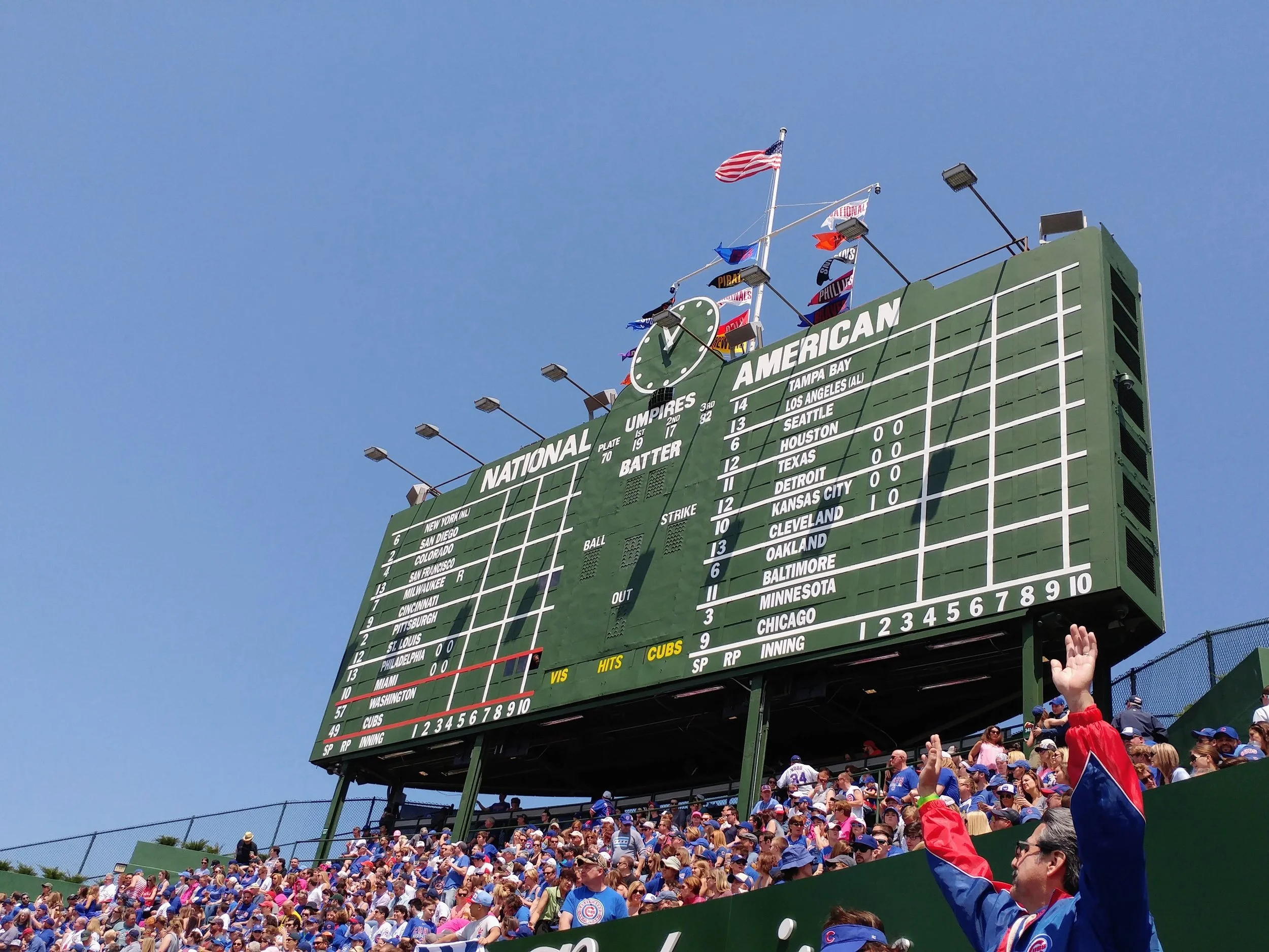 Crowd of baseball fans at a stadium, cheering and waving flags, with a large scoreboard in the background displaying game scores and team names, including the Chicago Cubs.