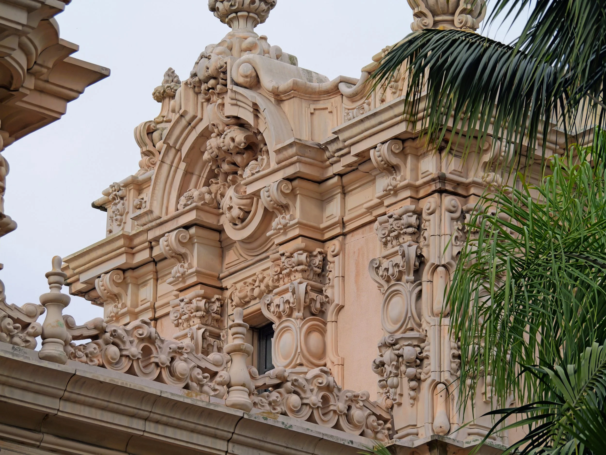 Close-up of an ornate beige stone building with detailed decorative carvings and a palm tree branch partially visible on the right.