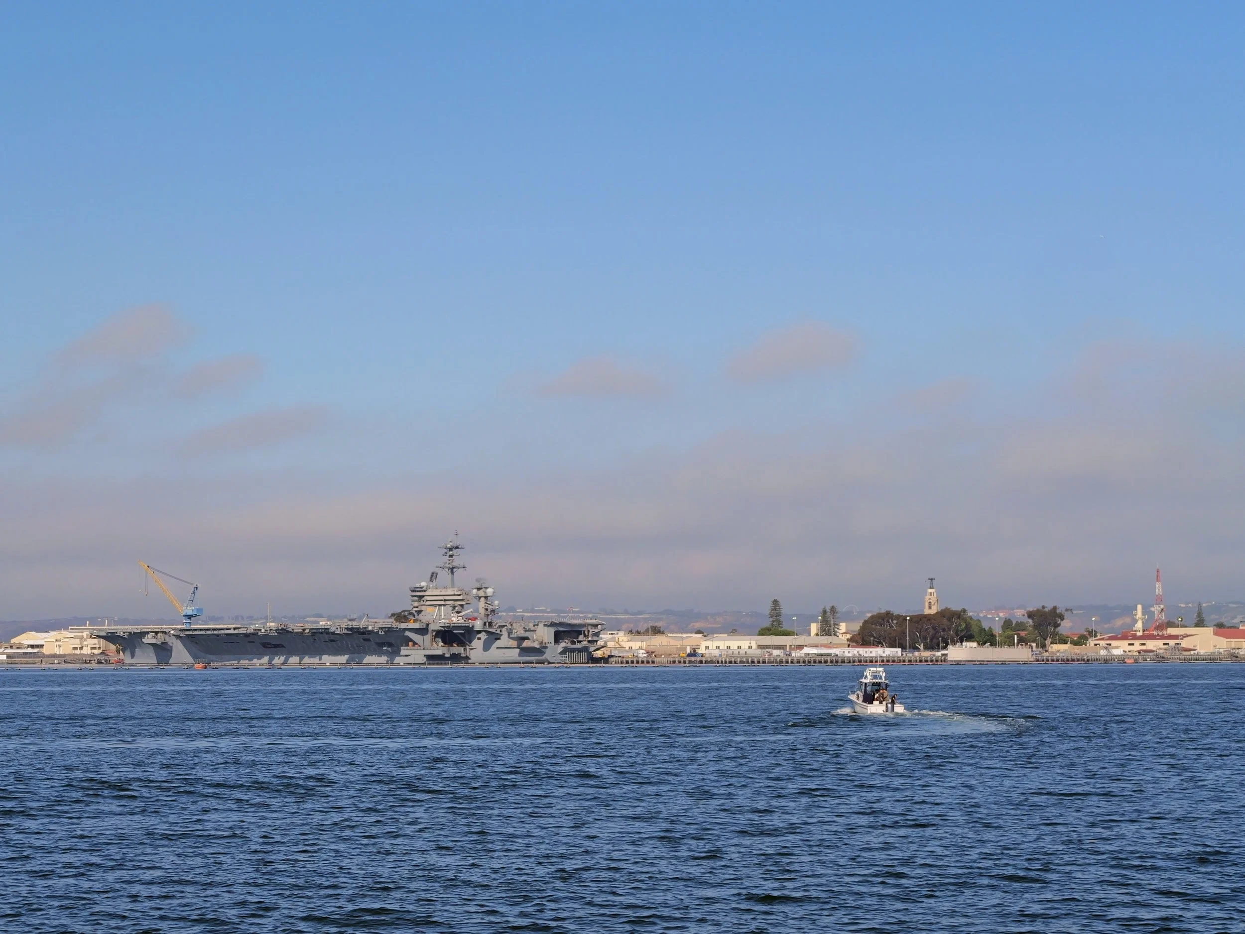 A large aircraft carrier ship on the water with a city skyline and blue sky in the background.