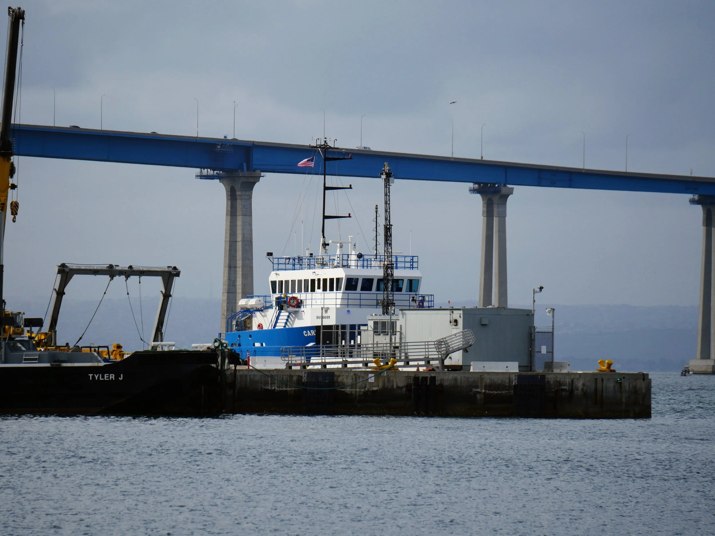 A dock with boats and a large blue bridge overhead, a crane, some equipment, and water in the foreground.