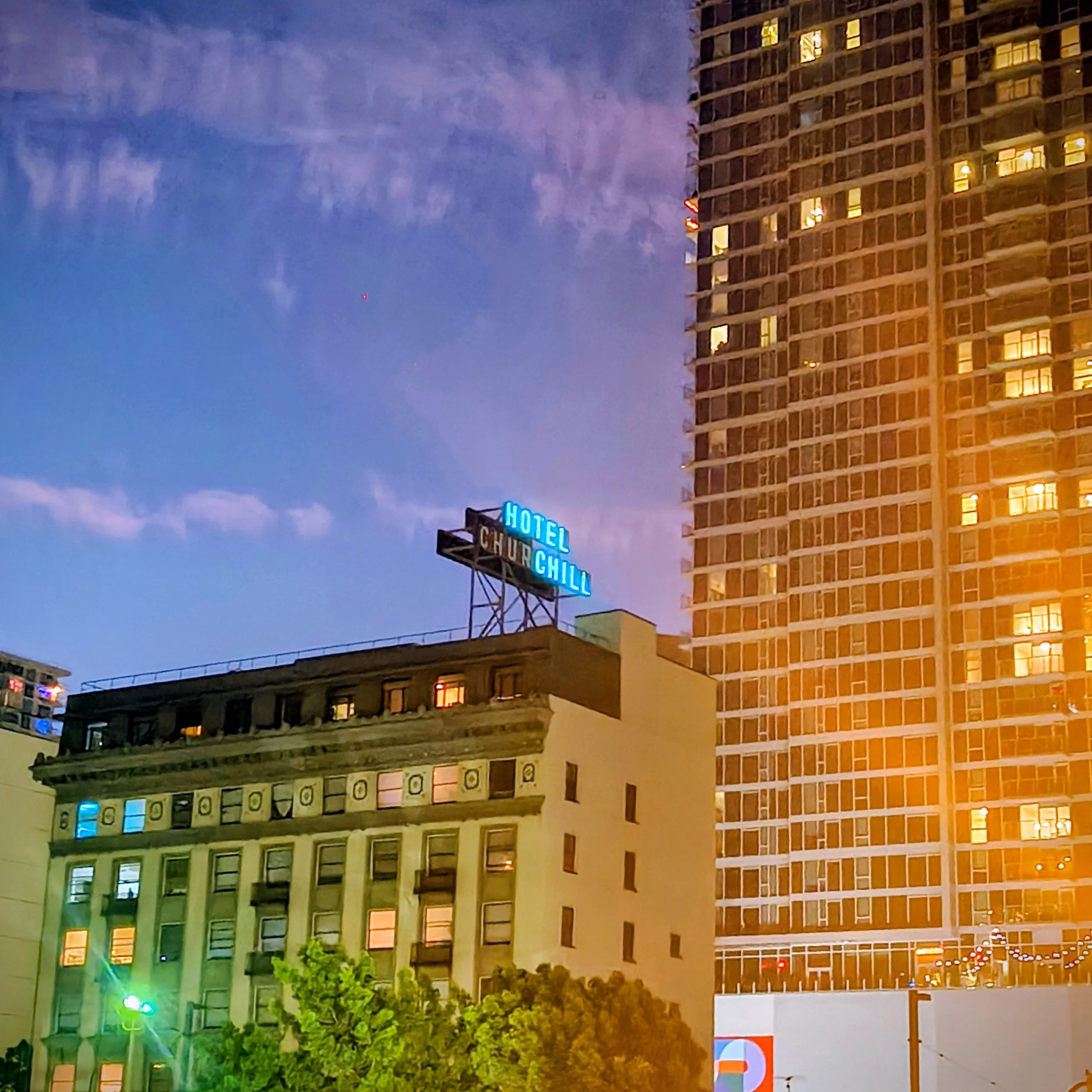 Nighttime city scene with a tall building featuring a blue neon sign that reads 'HOTEL CHILLI' and a large, modern high-rise building with illuminated windows.