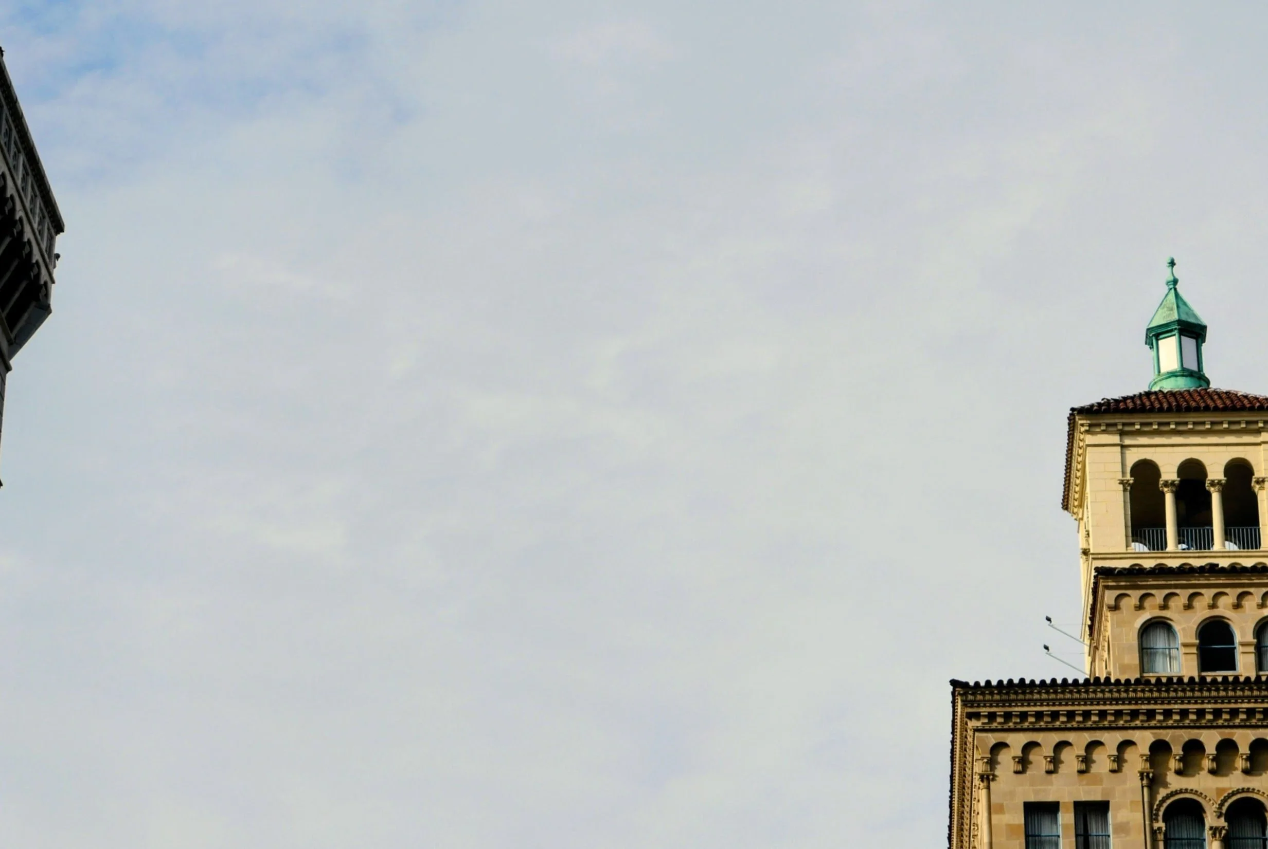 A tall beige building with arched windows and a green spire on top, seen against a cloudy sky. Part of another building is visible on the left side of the image.