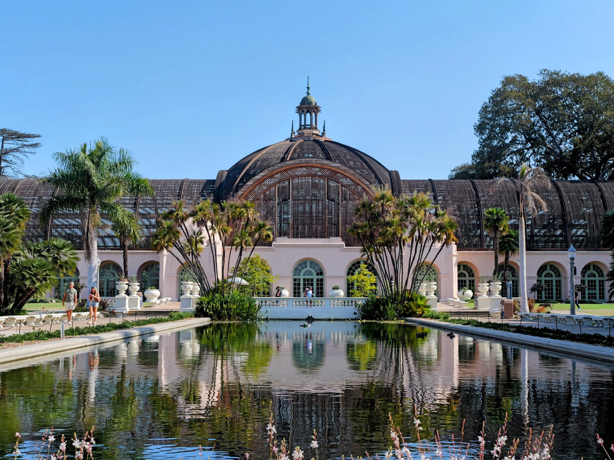 A historic building with a domed roof and ornate architectural features, surrounded by palm trees and a reflecting pond in the foreground under a clear blue sky.
