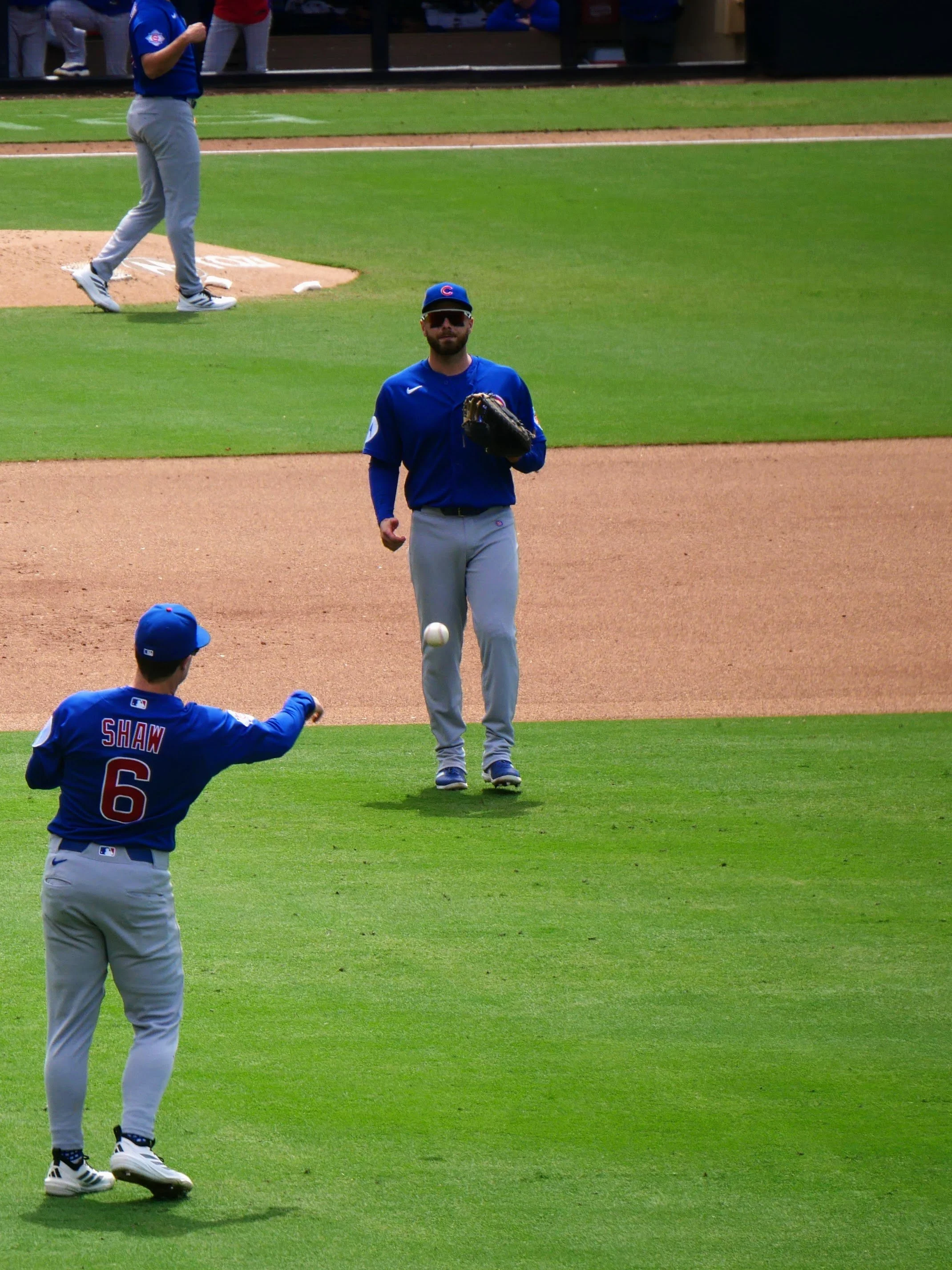 Chicago Cubs baseball players on the field during a game, one player jogging towards the camera, another preparing to catch the ball, with a player in the outfield wearing a cap with "Shaw 6" on the back.