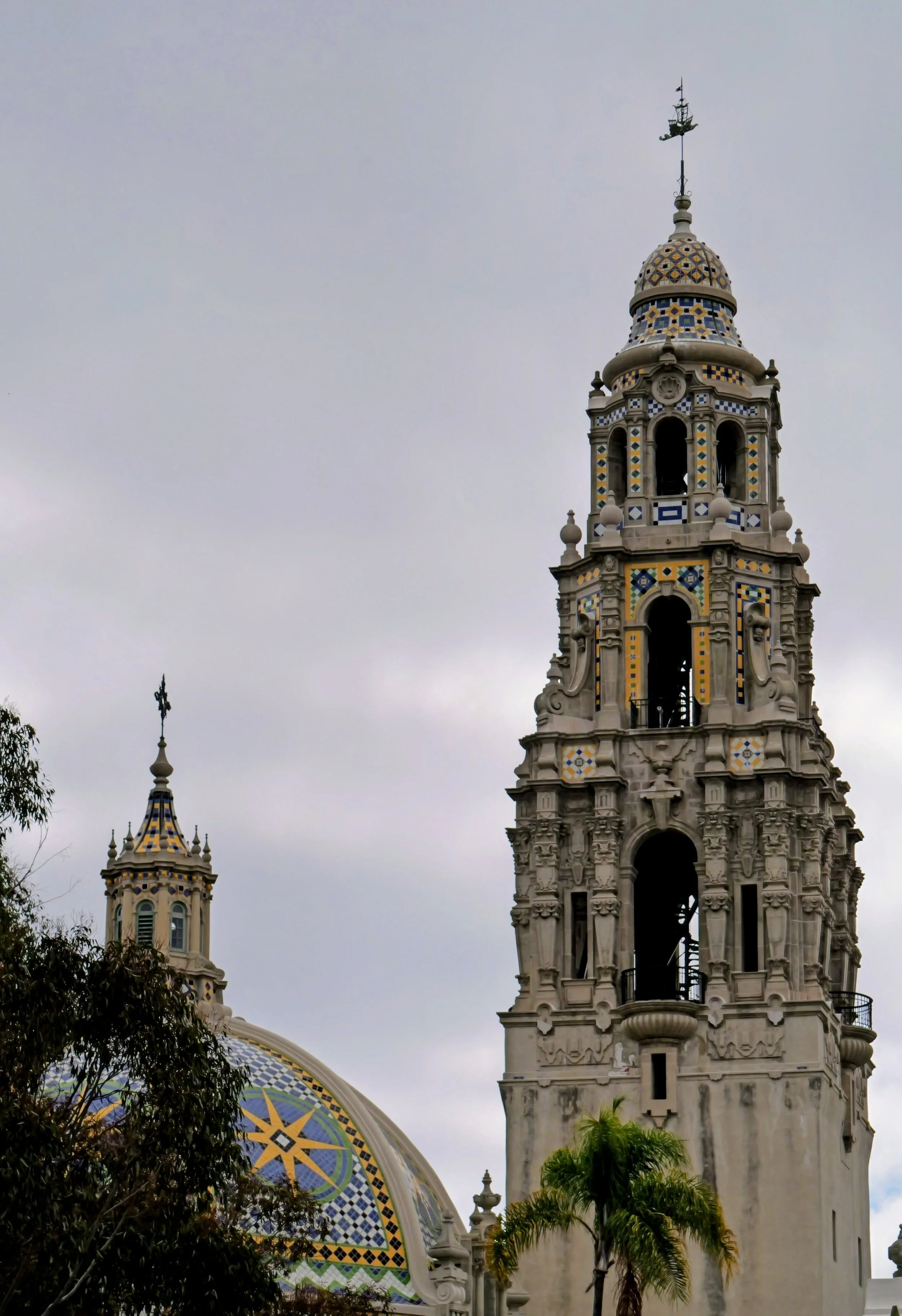 Historical building with ornate architecture and a colorful tile dome, with a cloudy sky in the background and trees in the foreground.