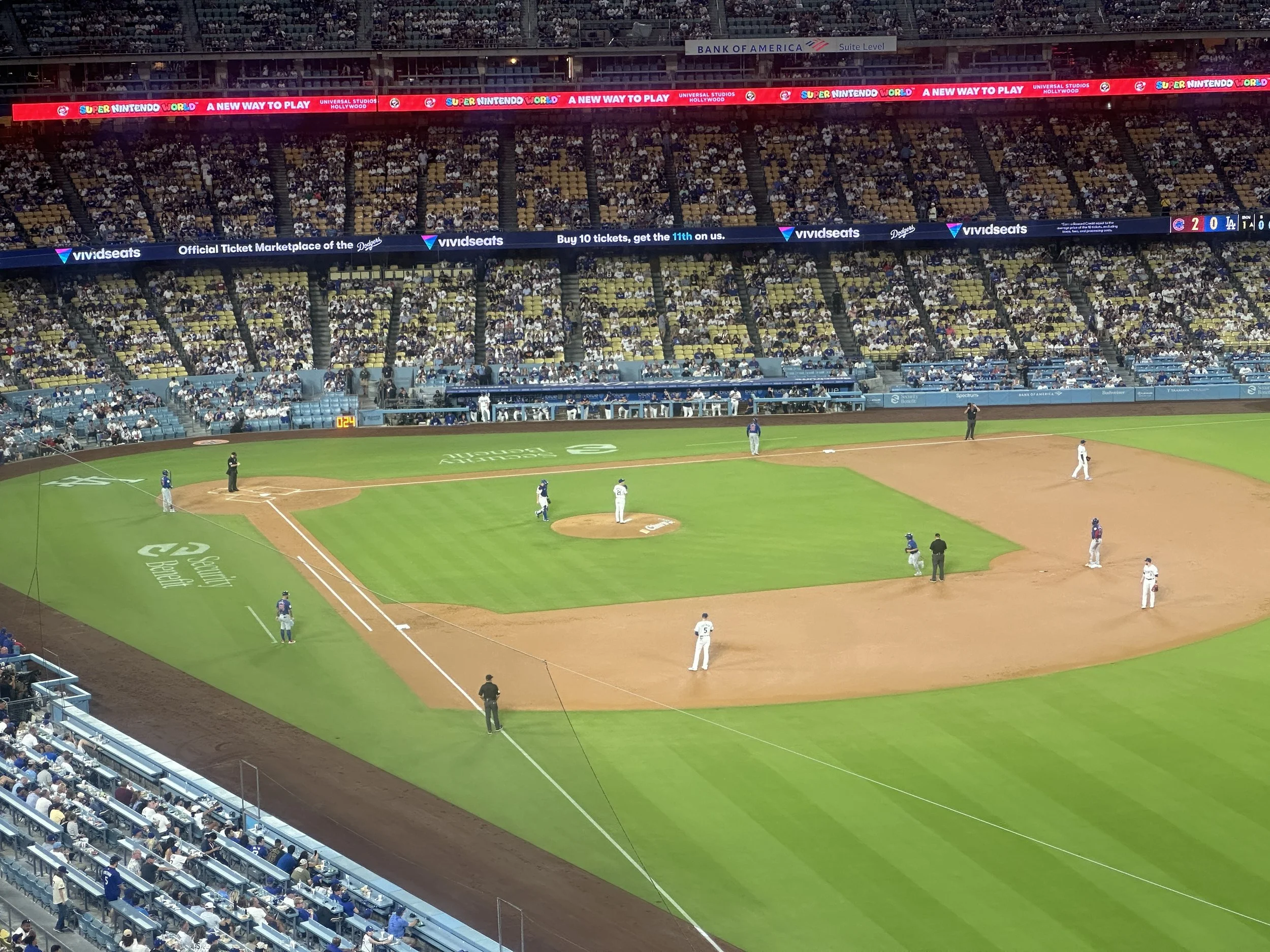 A baseball stadium with spectators in the stands, and players on the field. There are digital advertisements and banners around the stadium, including one for "Super Nintendo World."