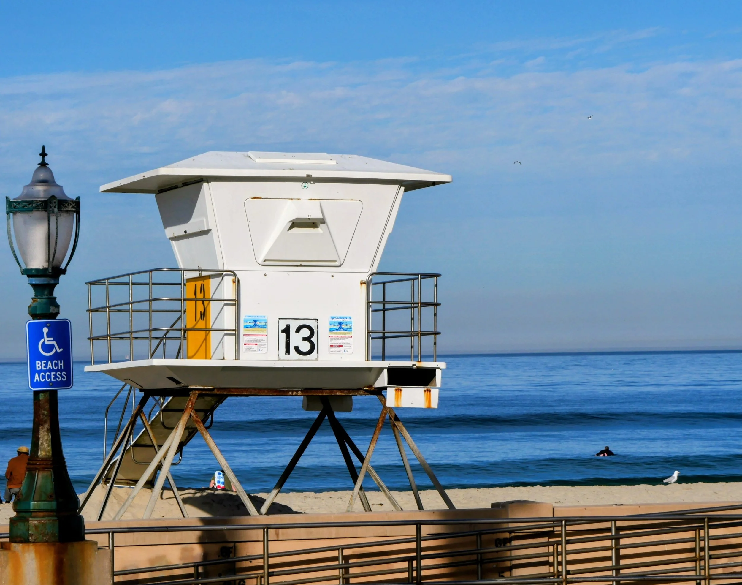 Lifeguard tower numbered 13 on a beach near the ocean with a blue sky and seagulls flying above. A person is swimming in the water, and a seagull is on the beach. There is a street lamp with a sign indicating beach access for handicapped.