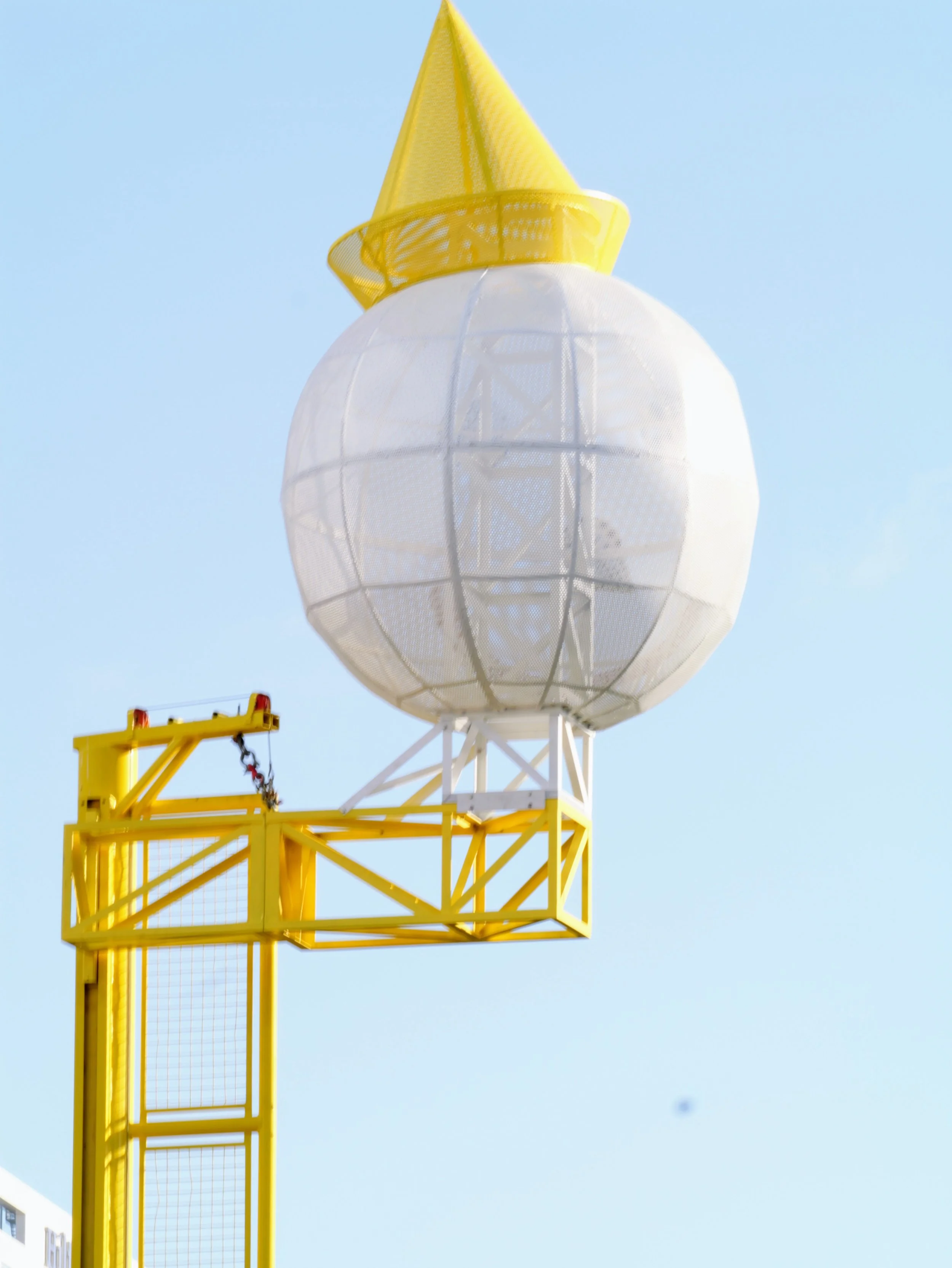 A spherical white structure elevated on a yellow lattice tower with a yellow cone-shaped top, set against a clear blue sky.