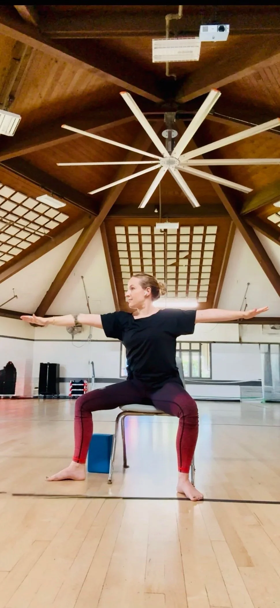 A woman in a black shirt and red leggings practicing yoga or stretching in a large indoor gymnasium with wooden ceiling beams, a ceiling fan, and a wooden floor.