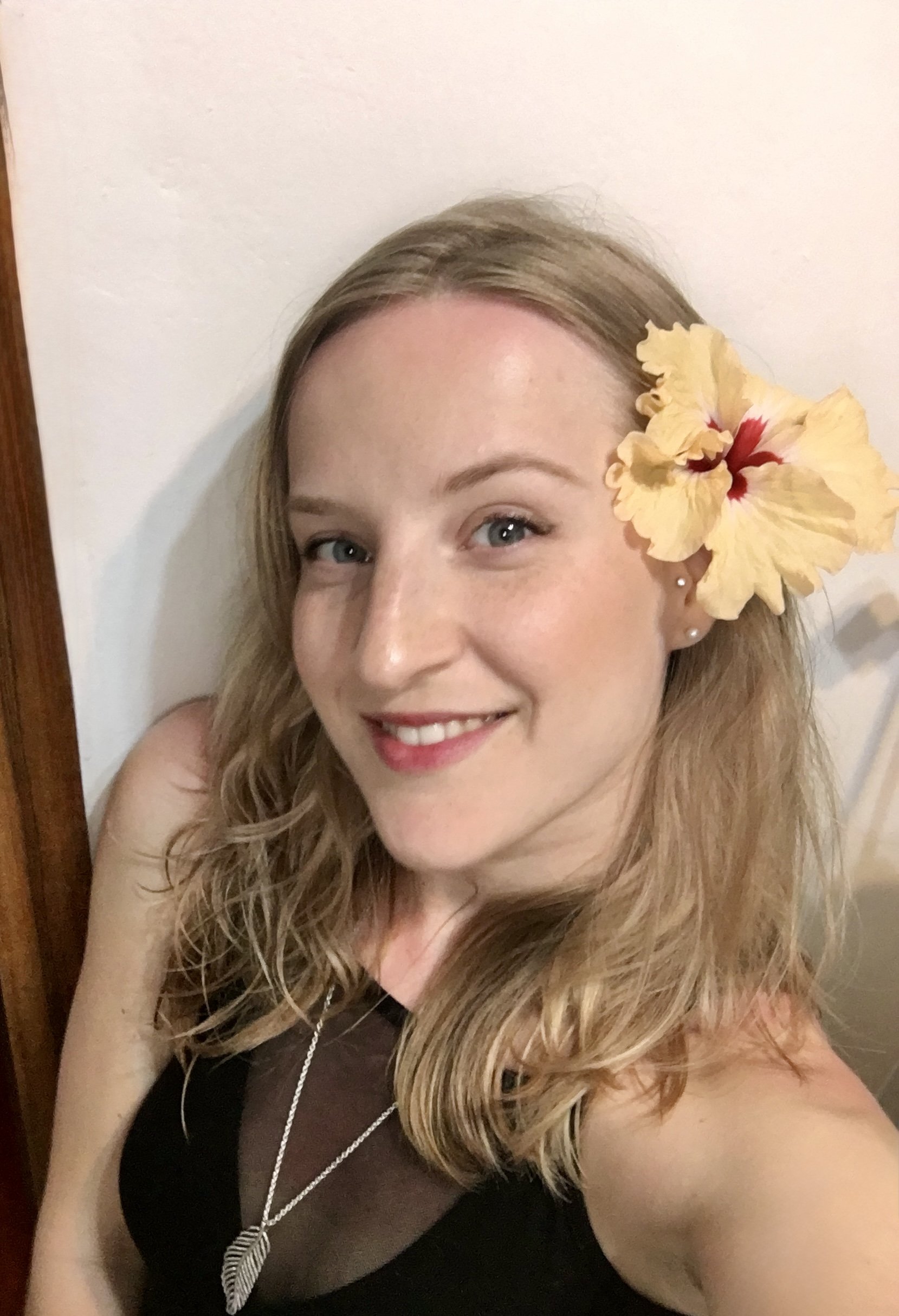 A smiling woman with blonde wavy hair, blue eyes, and a large yellow hibiscus flower in her hair, wearing a black top and a necklace with a feather pendant, standing against a white wall.