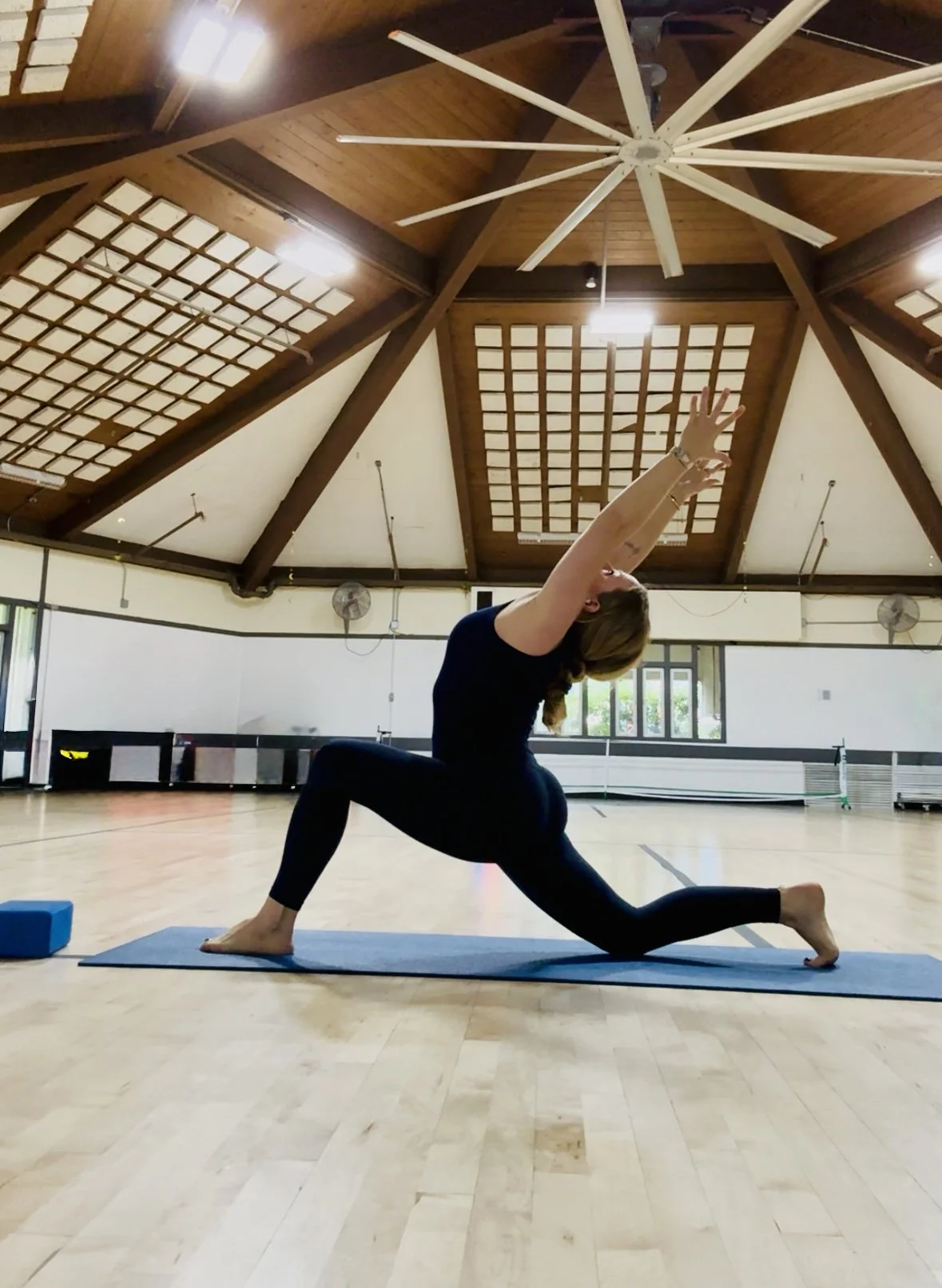 A woman practicing yoga in a spacious indoor studio with a wooden ceiling and large windows, performing a lunge pose with arms reaching upward.