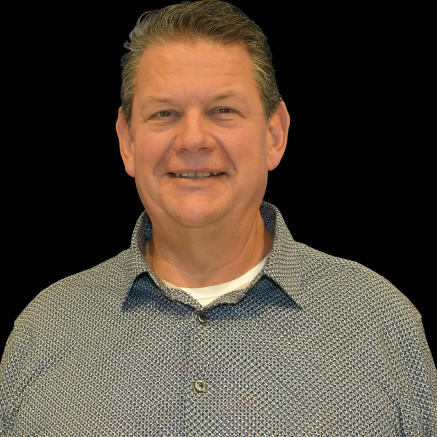 Black and white portrait of a middle-aged man smiling, wearing a patterned shirt, against a dark background.