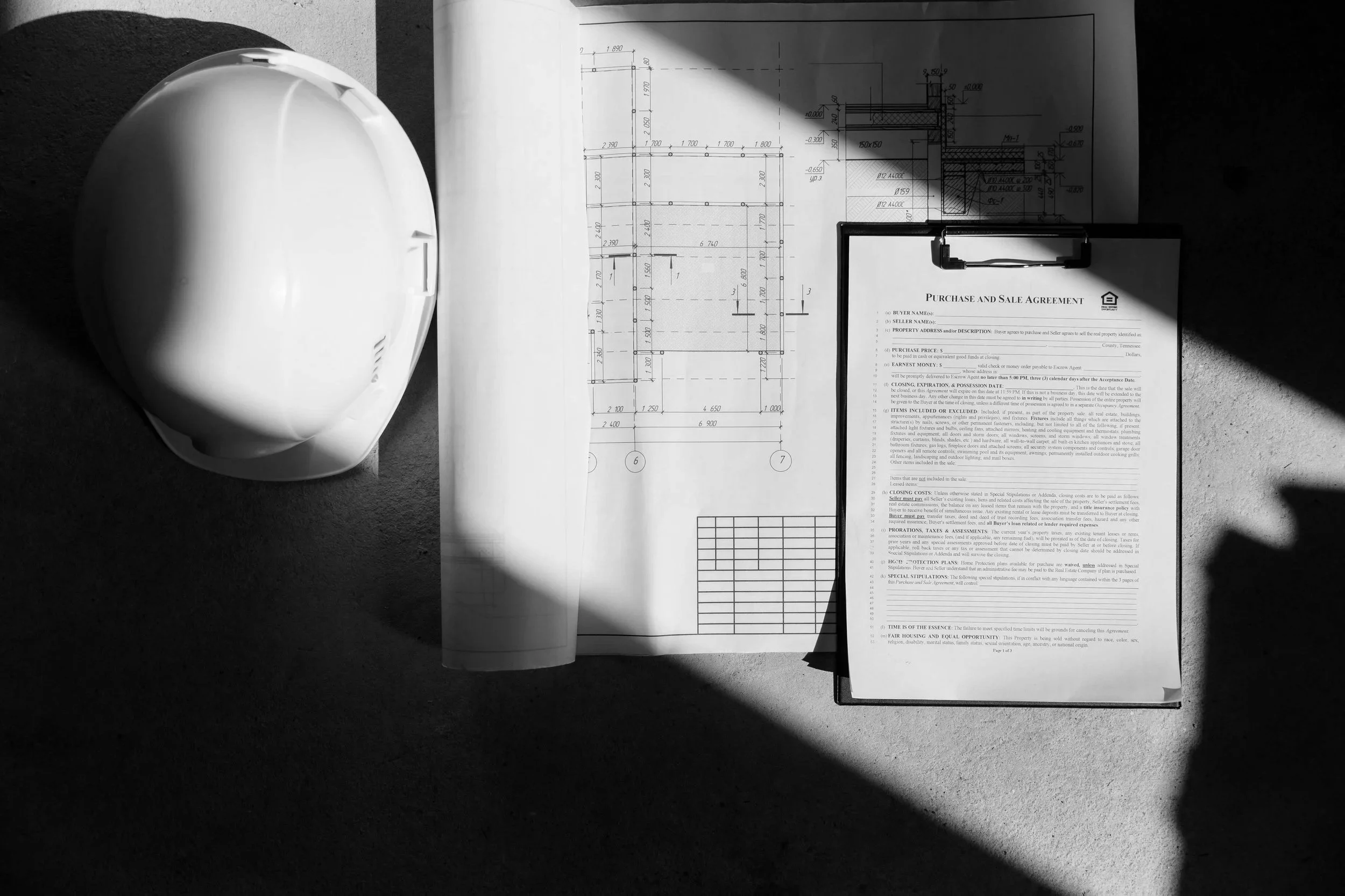Construction helmet, architectural blueprint, and contract on clipboard on a concrete surface, with a shadow cast across the scene.