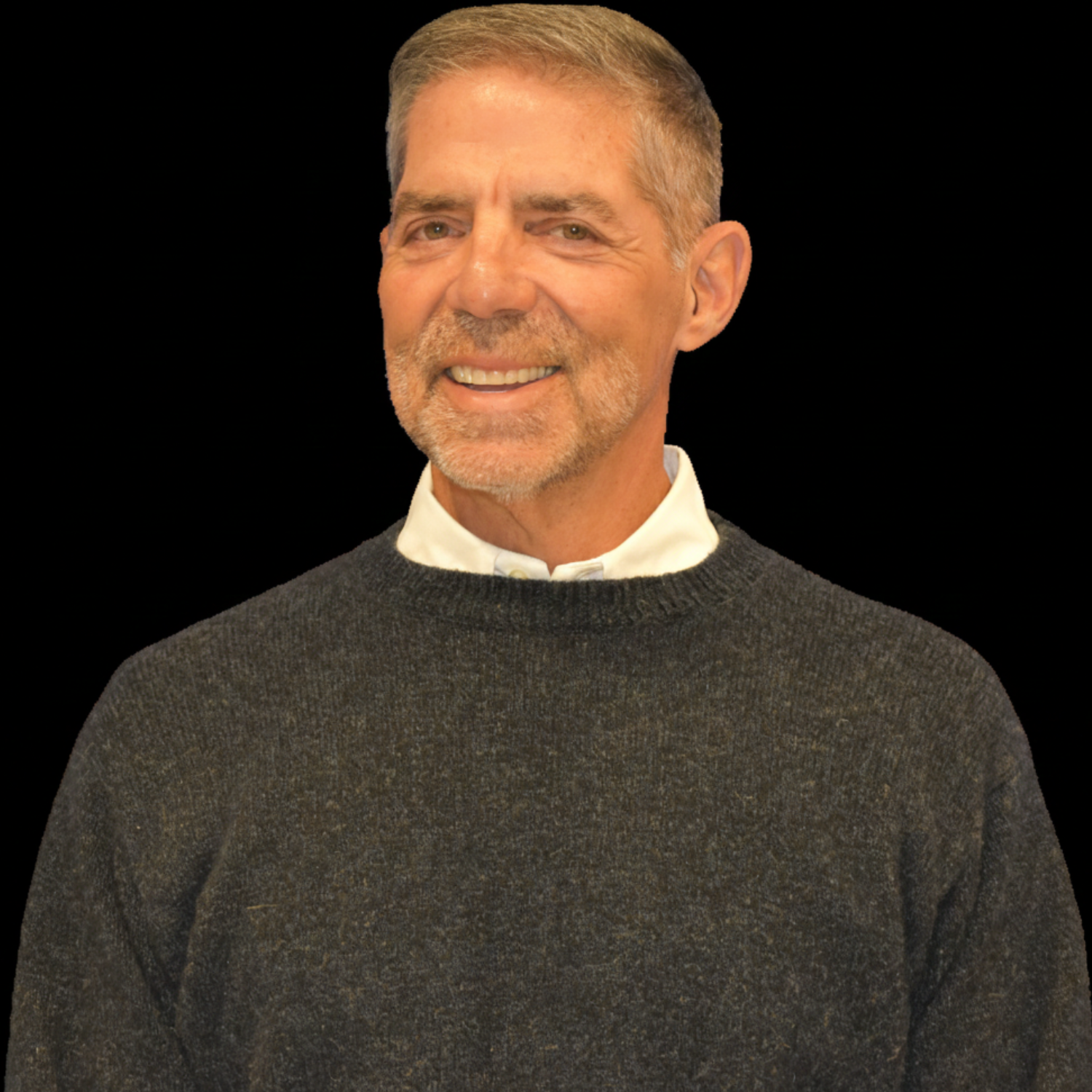 A black-and-white portrait of a smiling middle-aged man with a beard and short hair, wearing a sweater over a collared shirt, against a solid black background.