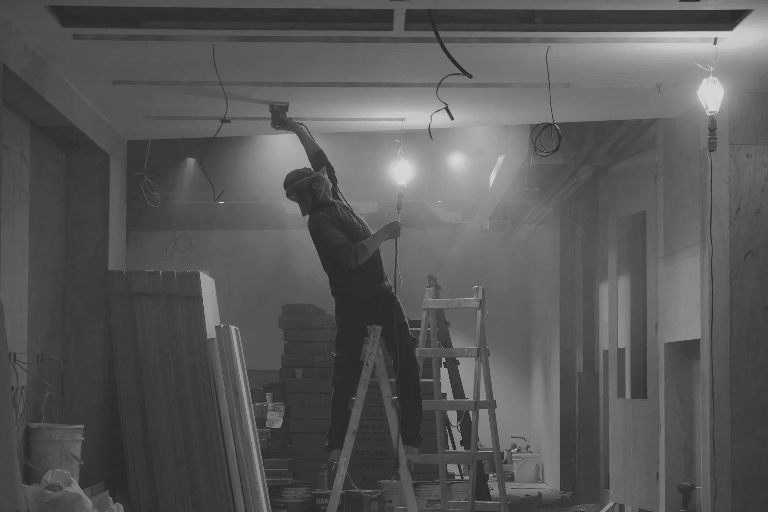 A worker in a face mask and safety glasses standing on a ladder, hanging wires from the ceiling of a construction site.