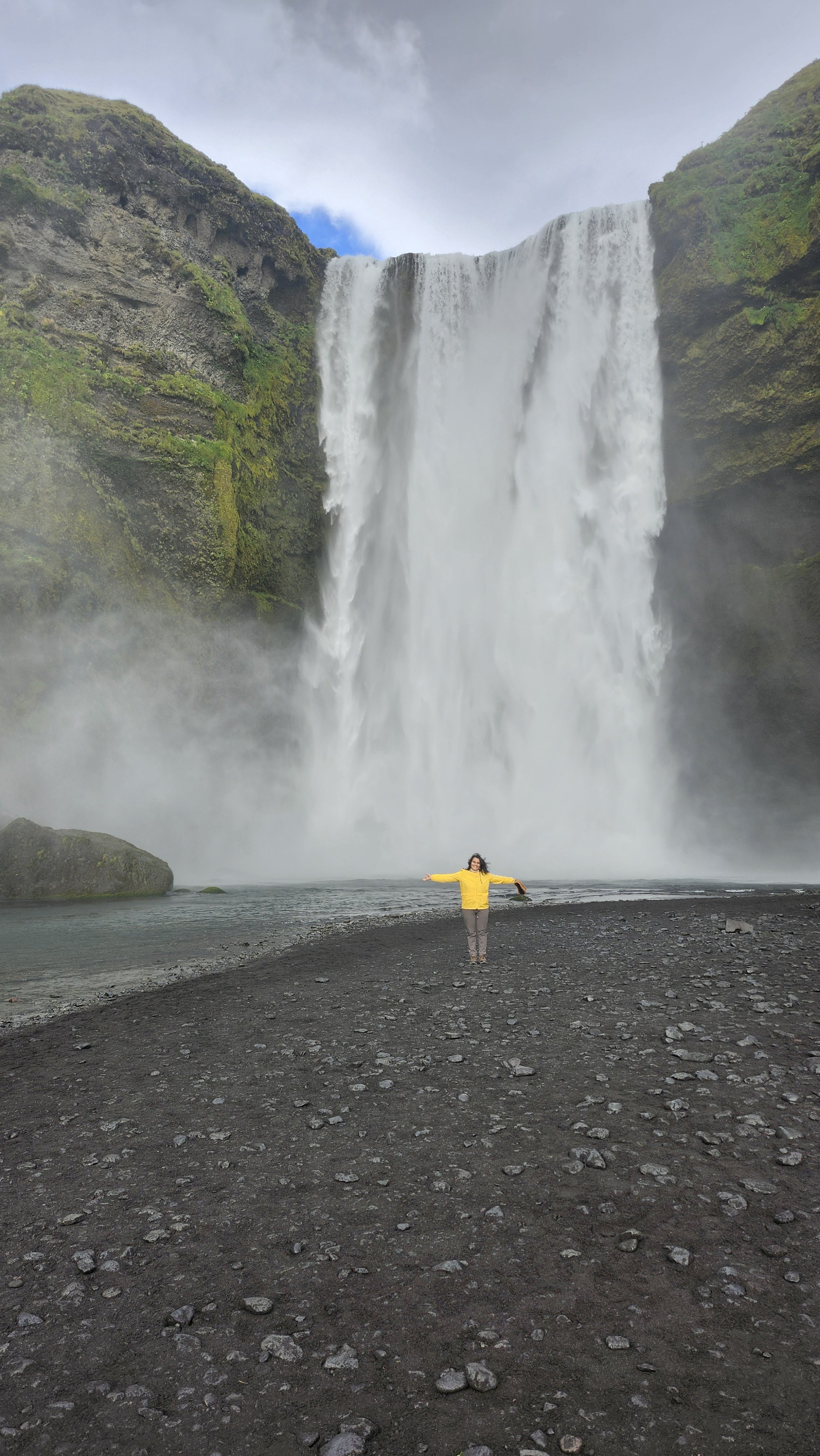 Person in yellow jacket standing with arms outstretched in front of a large waterfall with mist, rocky black beach, and moss-covered cliffs.