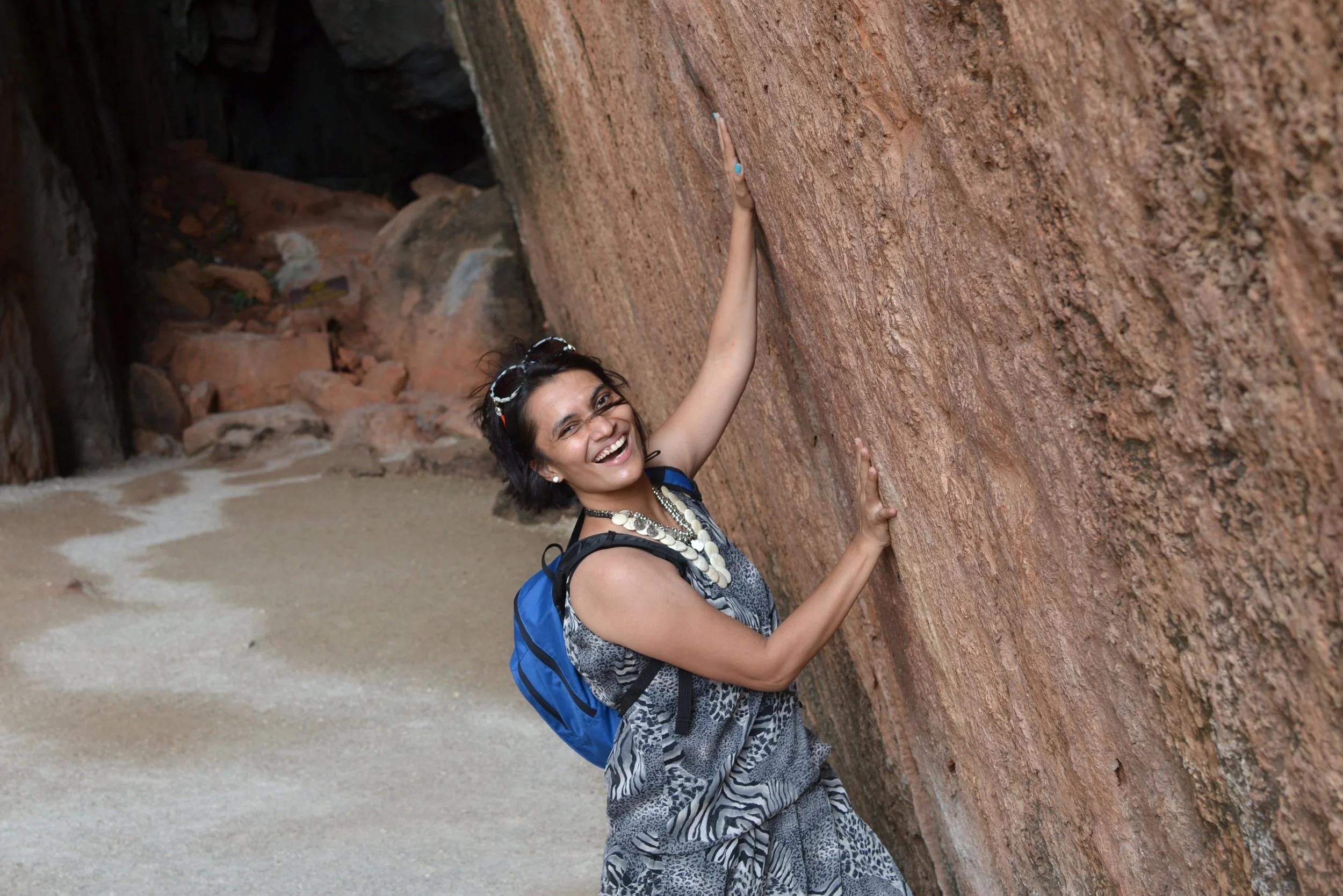 Woman smiling and climbing a large red rock formation on a sandy beach, wearing a patterned dress, glasses on her head, and a blue backpack.