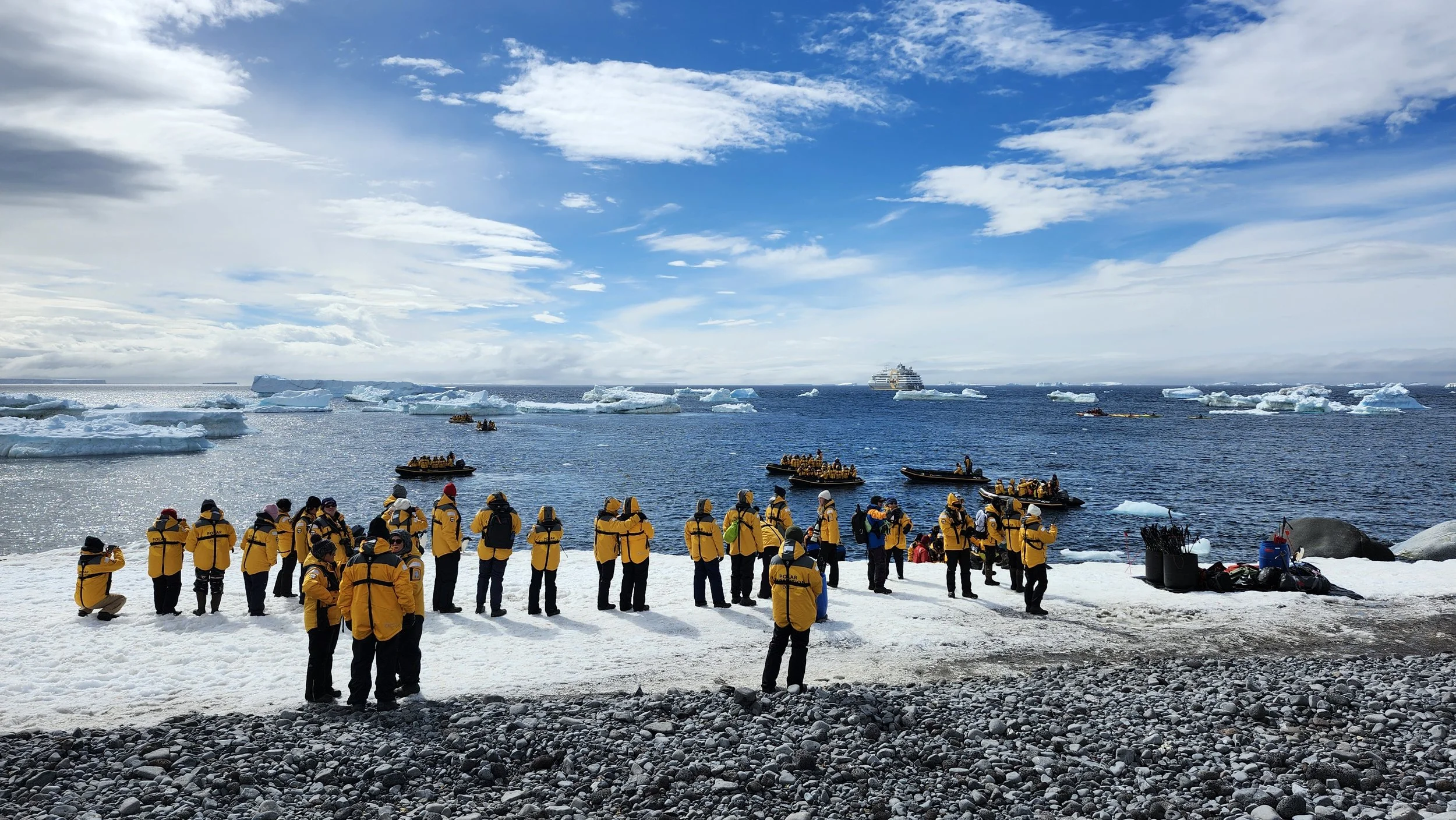 A group of Quark expeiditon tourists a snowy shore in Antarctica, observing icebergs and the Ultramarine cruise ship in the icy water.