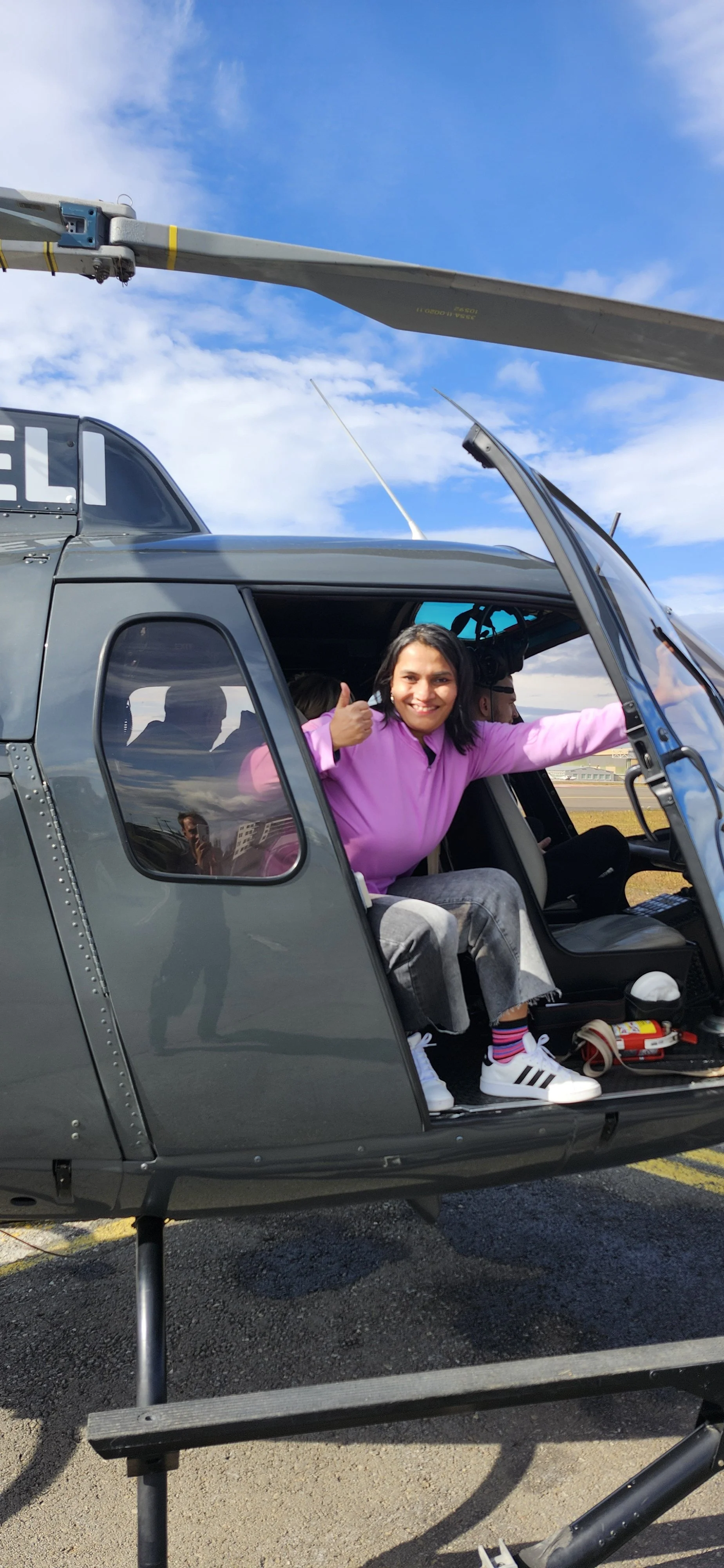 Woman in pink jacket sitting in helicopter cockpit, smiling and giving a thumbs-up, with the open door, blue sky, and pilot visible.