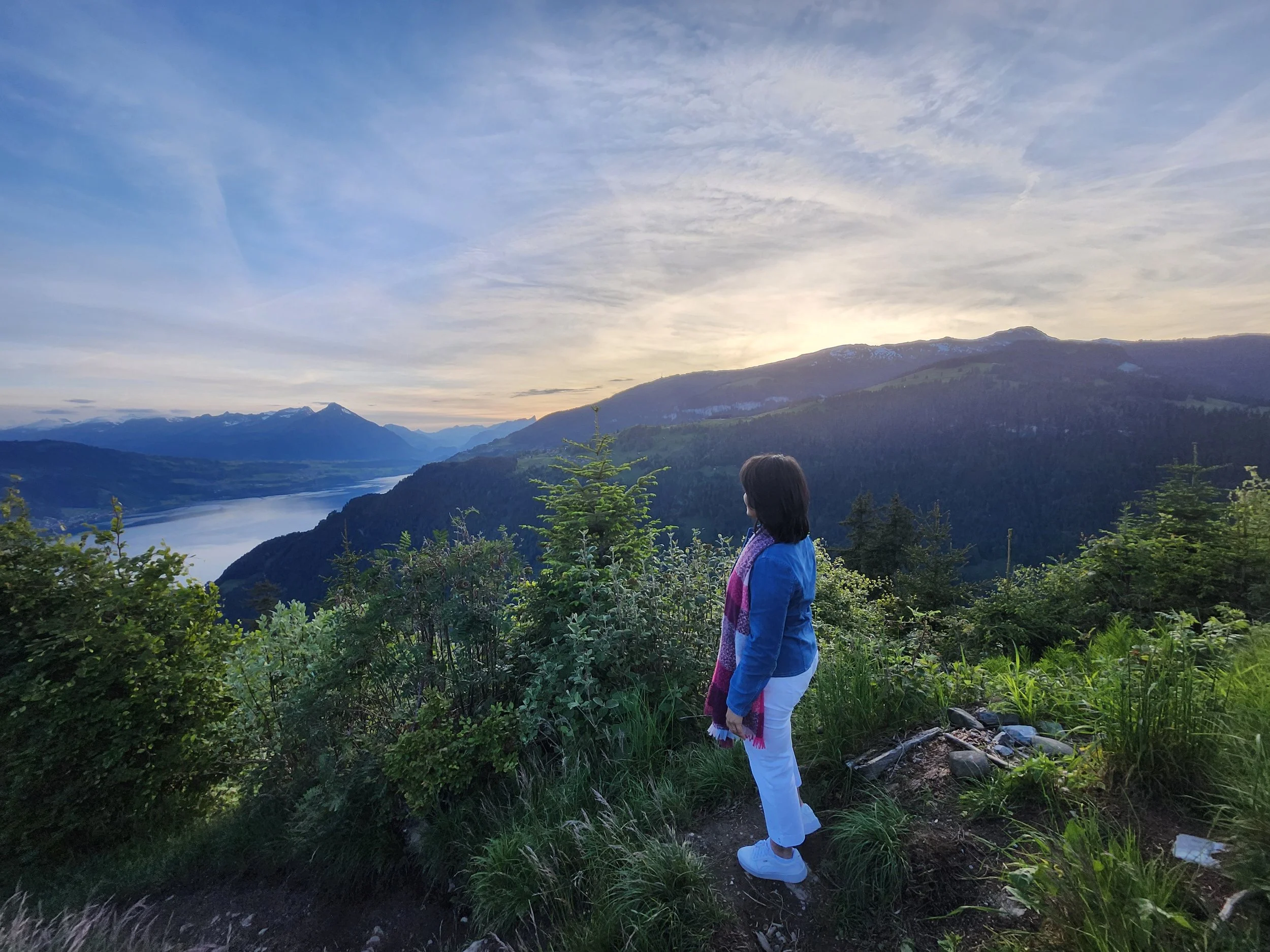 A woman standing on a mountain trail, looking over a scenic river and mountain landscape during sunset.