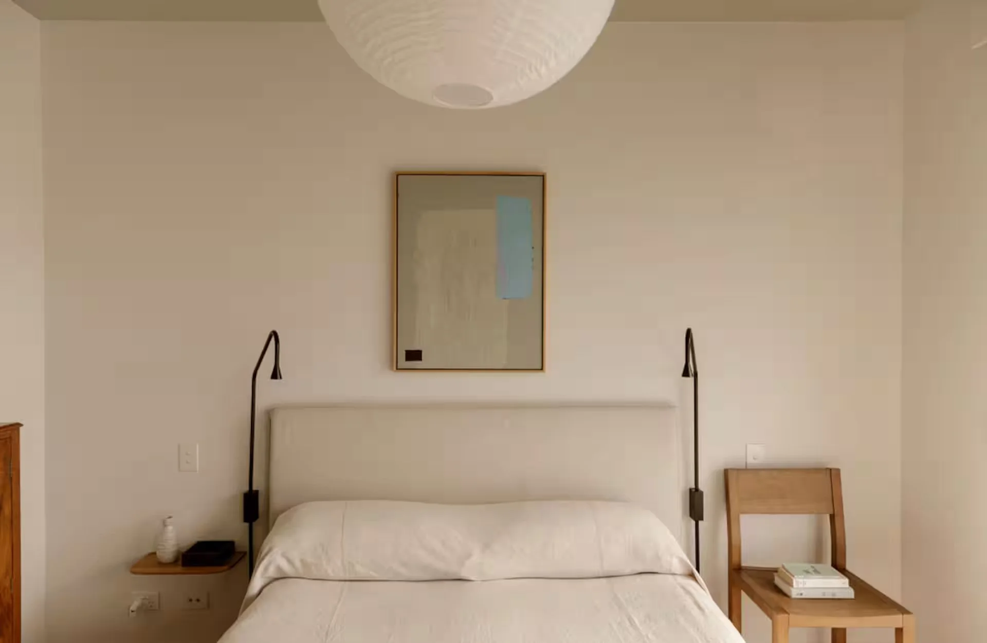 A minimalist bedroom featuring a white bed with a white headboard, black wall-mounted reading lamps on each side, a small wooden side table with a white bottle and black tray, a wooden chair with stacked books, and a framed mirror hanging above the bed. A paper lantern ceiling light hangs from the ceiling.