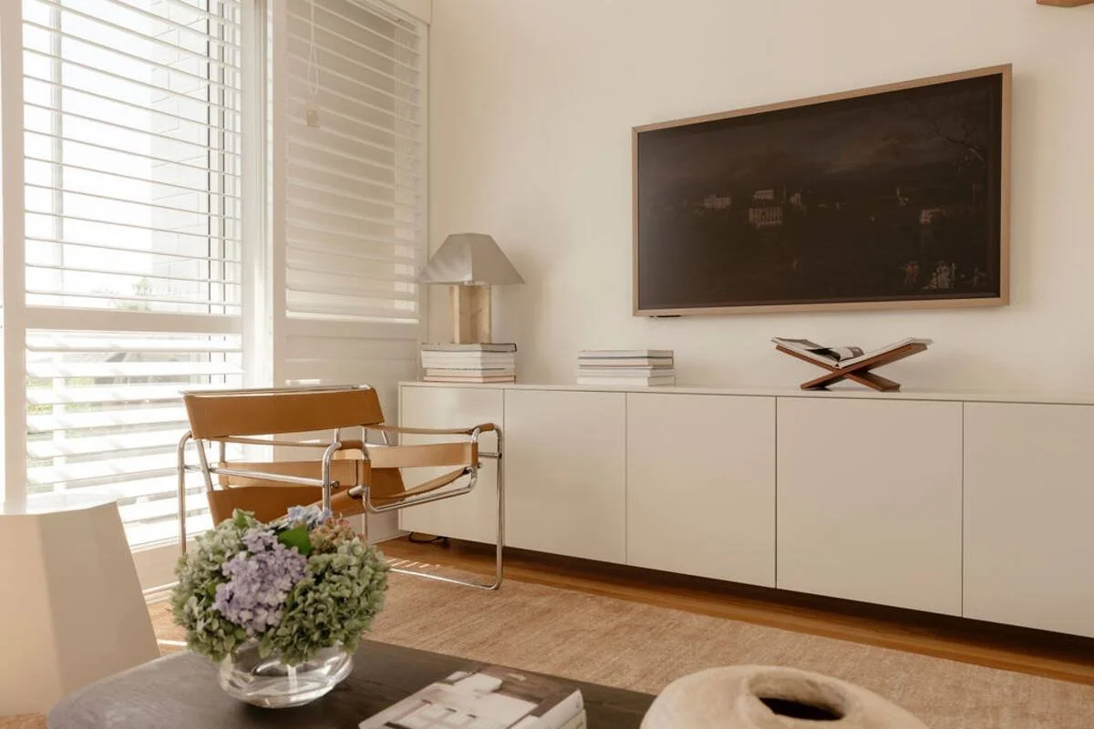 Bright living room with a large window, a modern leather chair, a side table with a lamp and books, a wall-mounted TV, a console with books and a magazine holder, and a flower arrangement on a dining table.