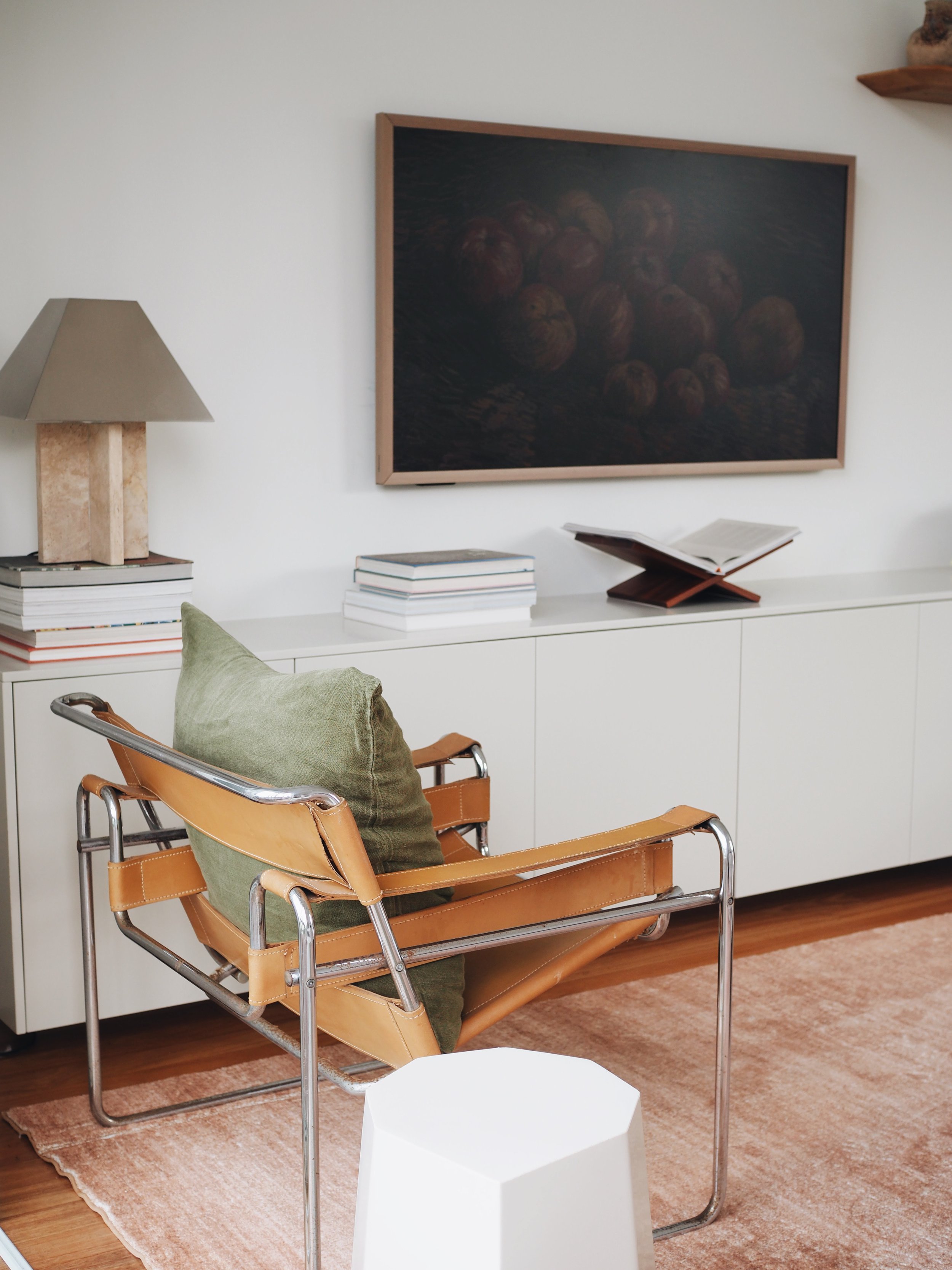 A cozy living room corner featuring a mid-century modern leather and chrome chair with a green cushion, a white sideboard topped with stacked books and an open book on a wooden stand, a table lamp, a large dark still life painting of apples on the wall, and a peach-colored area rug.