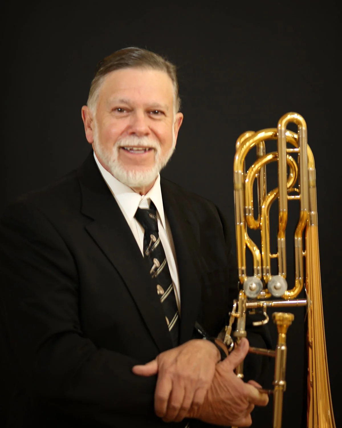 A smiling man in a black suit holding a gold trombone against a black background.