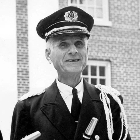 An elderly man dressed in a formal military or police uniform with a cap, standing outdoors in front of a brick building.