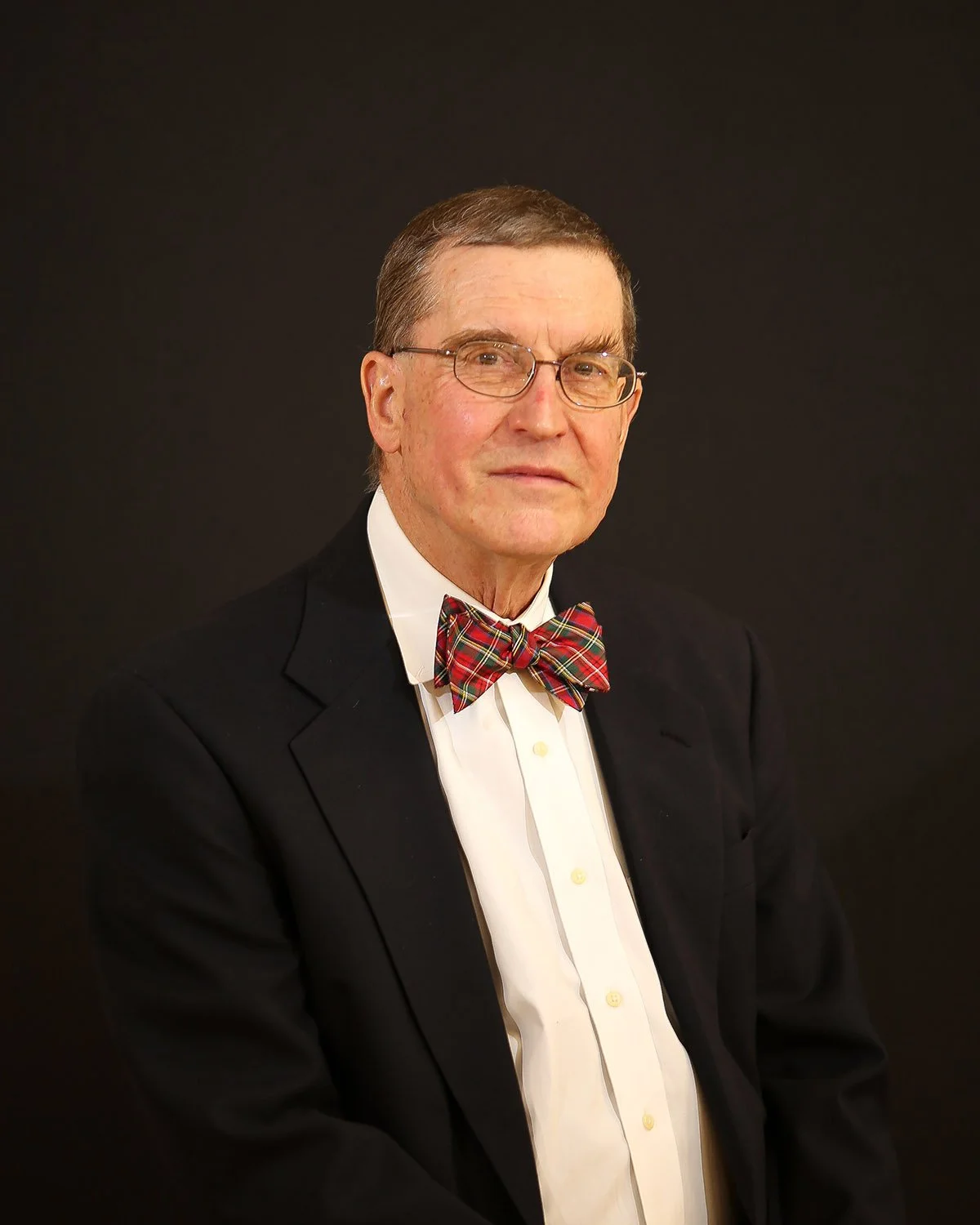 Portrait of an elderly man in a tuxedo with a plaid bow tie and glasses, against a dark background.