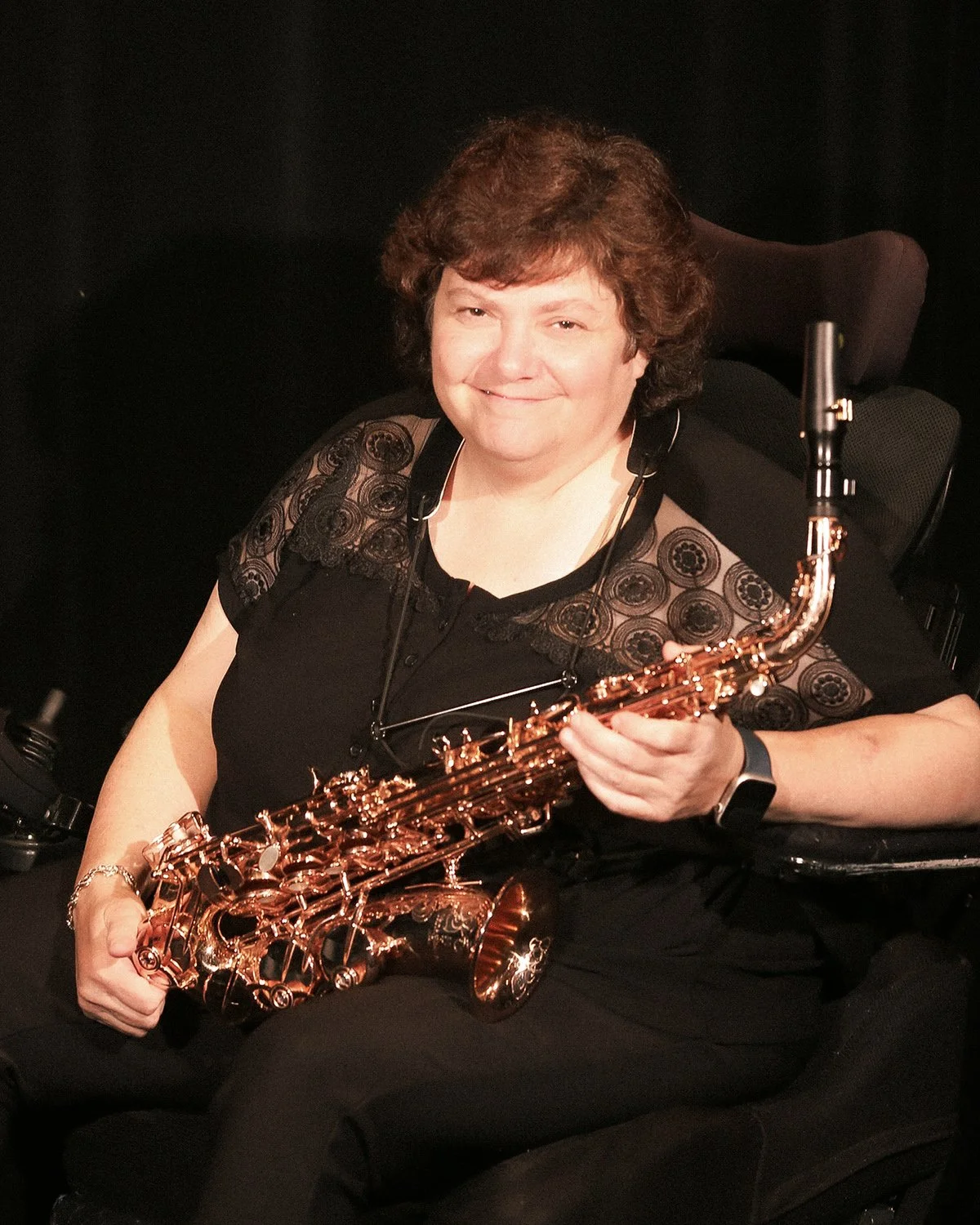 Woman with short curly hair smiling, seated in a motorized wheelchair, holding a shiny copper-colored saxophone, wearing a black top with lace details.