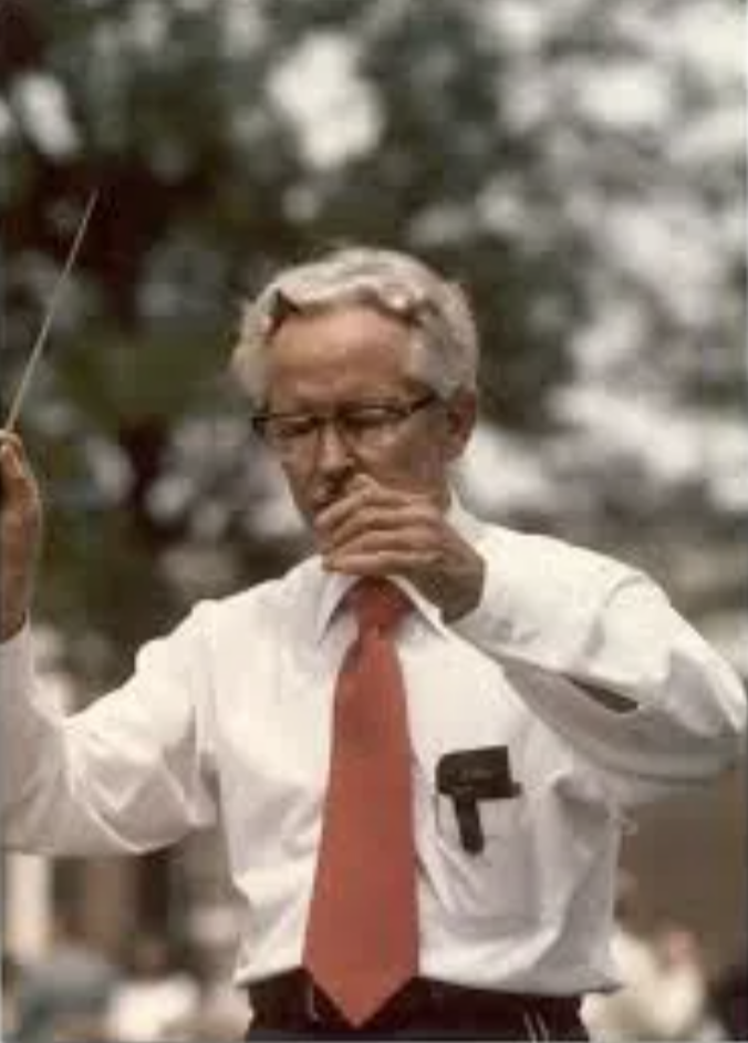 Older man with gray hair and glasses conducting with a baton, wearing a white dress shirt, red tie, and a black device clipped to his pocket, outdoors with trees in the background.