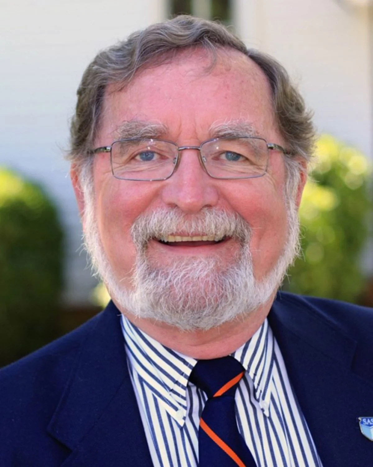 A smiling elderly man with glasses and a white beard, wearing a dark suit, striped shirt, and tie, outdoors with greenery background.