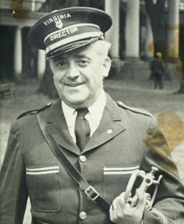 Black and white photo of a man in a police uniform, smiling, holding a device in his right hand, wearing a peaked cap labeled "Virginia" with the word "Director" on it, and a shoulder strap across his chest.