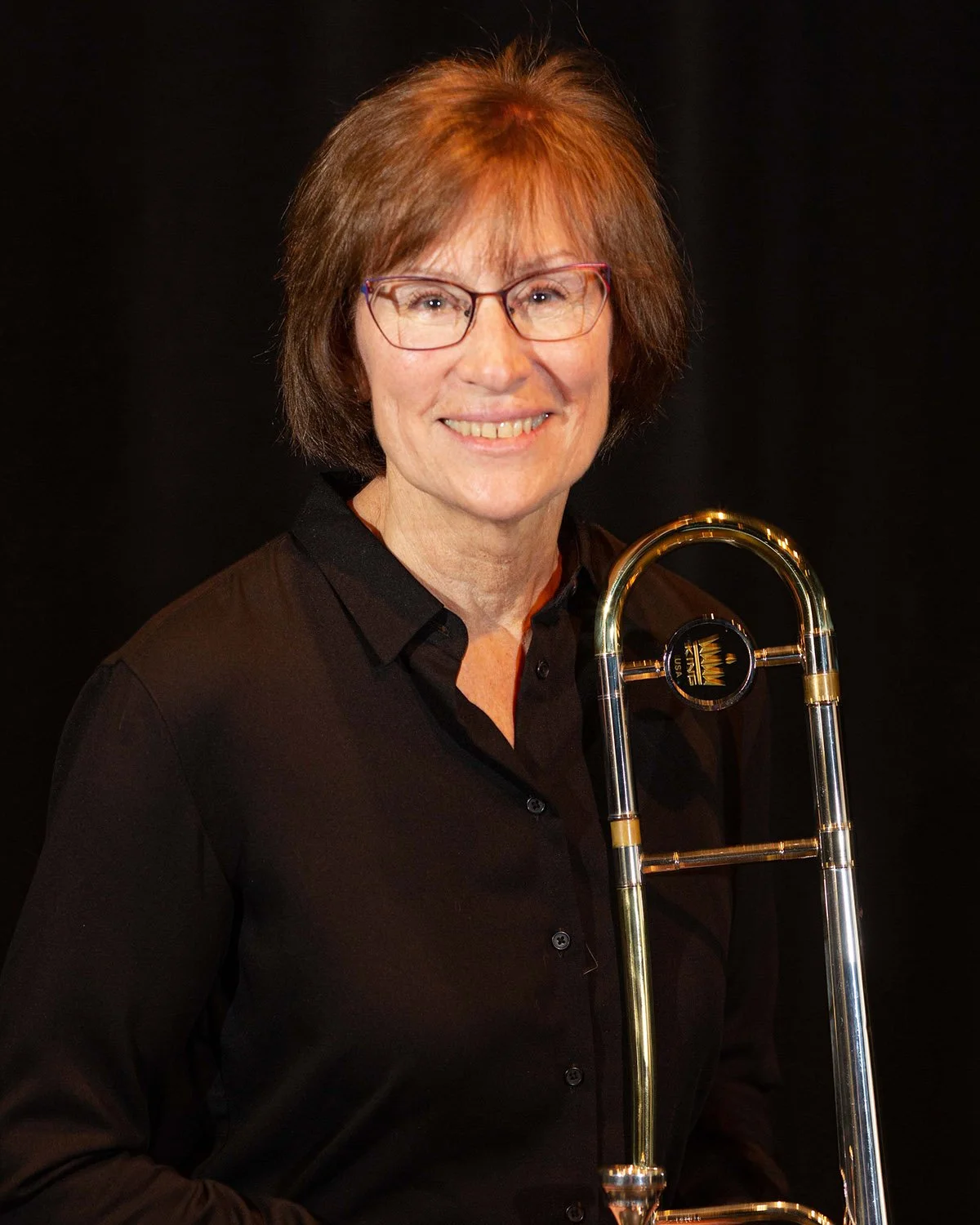 A smiling woman with glasses holding a trombone against a black background.