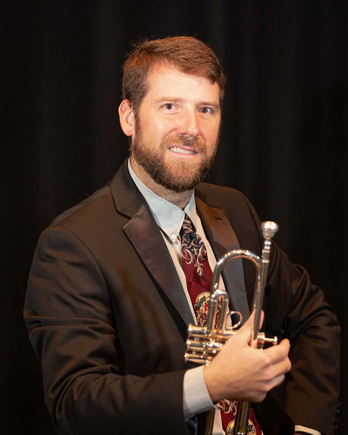 A man with a beard in a suit holding a trumpet, smiling at the camera.