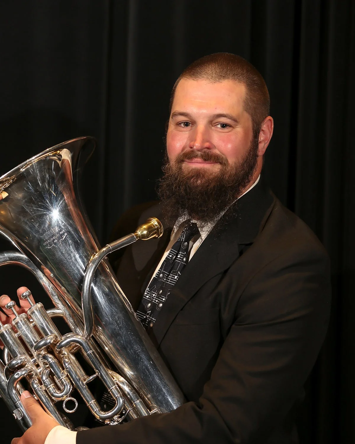 A man with a beard and short hair holding a brass musical instrument, possibly a euphonium or tuba, in front of a black curtain.