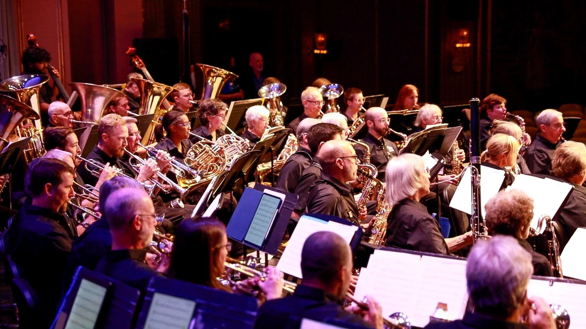 An orchestral band performing on stage, with musicians playing brass and woodwind instruments, reading sheet music, in a concert hall.