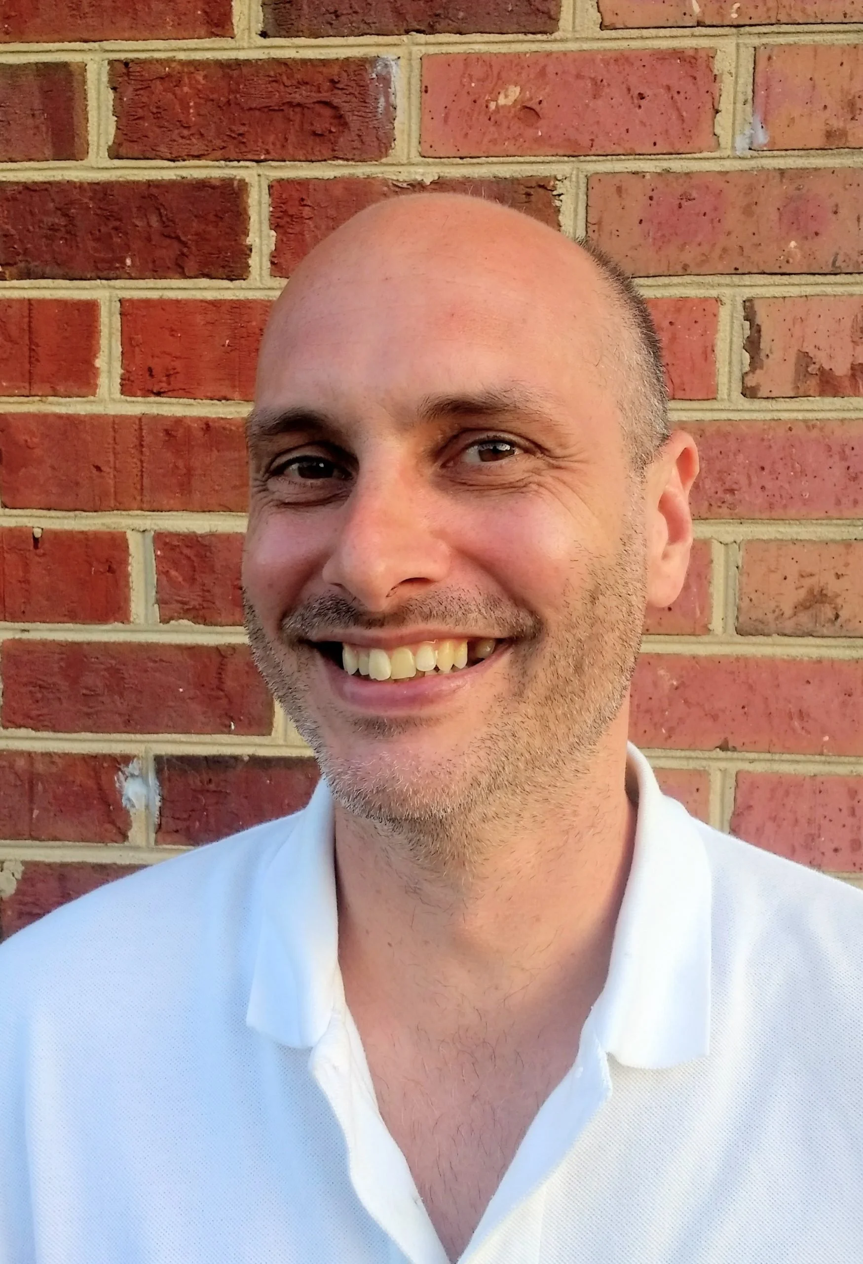 Close-up of a smiling man with a bald head and light beard, wearing a white shirt, standing against a brick wall.