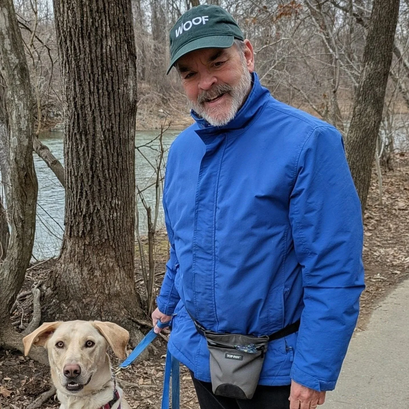 A man with a beard and gray hair wearing a blue jacket and a black cap with "WOOF" written on it, standing outdoors next to a tree near a river, holding a leash connected to a light-colored dog with floppy ears.