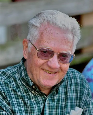 An elderly man with white hair, wearing glasses and a green checkered shirt, smiling outdoors.