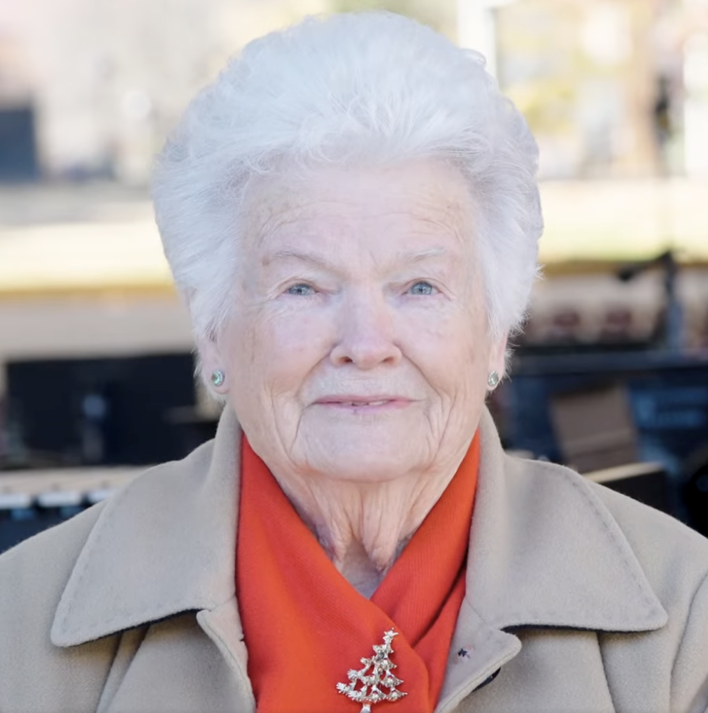 An elderly woman with white hair, wearing a beige coat, a red scarf with a Christmas tree brooch, and earrings. She is outdoors, with a blurred background of trees and benches.