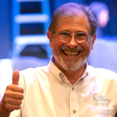 A smiling man with glasses and gray hair giving a thumbs-up gesture, wearing a white shirt with a logo.
