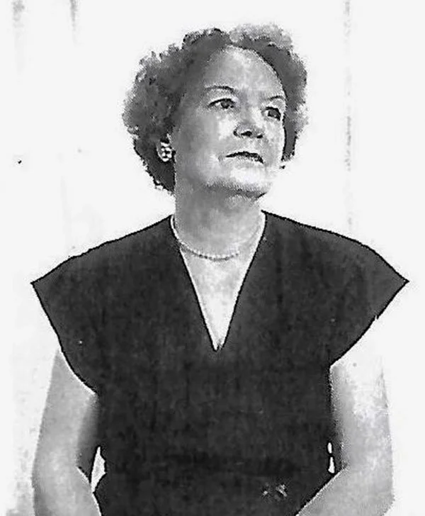 Black and white photo of a woman with short curly hair wearing a dark top, pearl necklace, and earrings, standing against a light background.