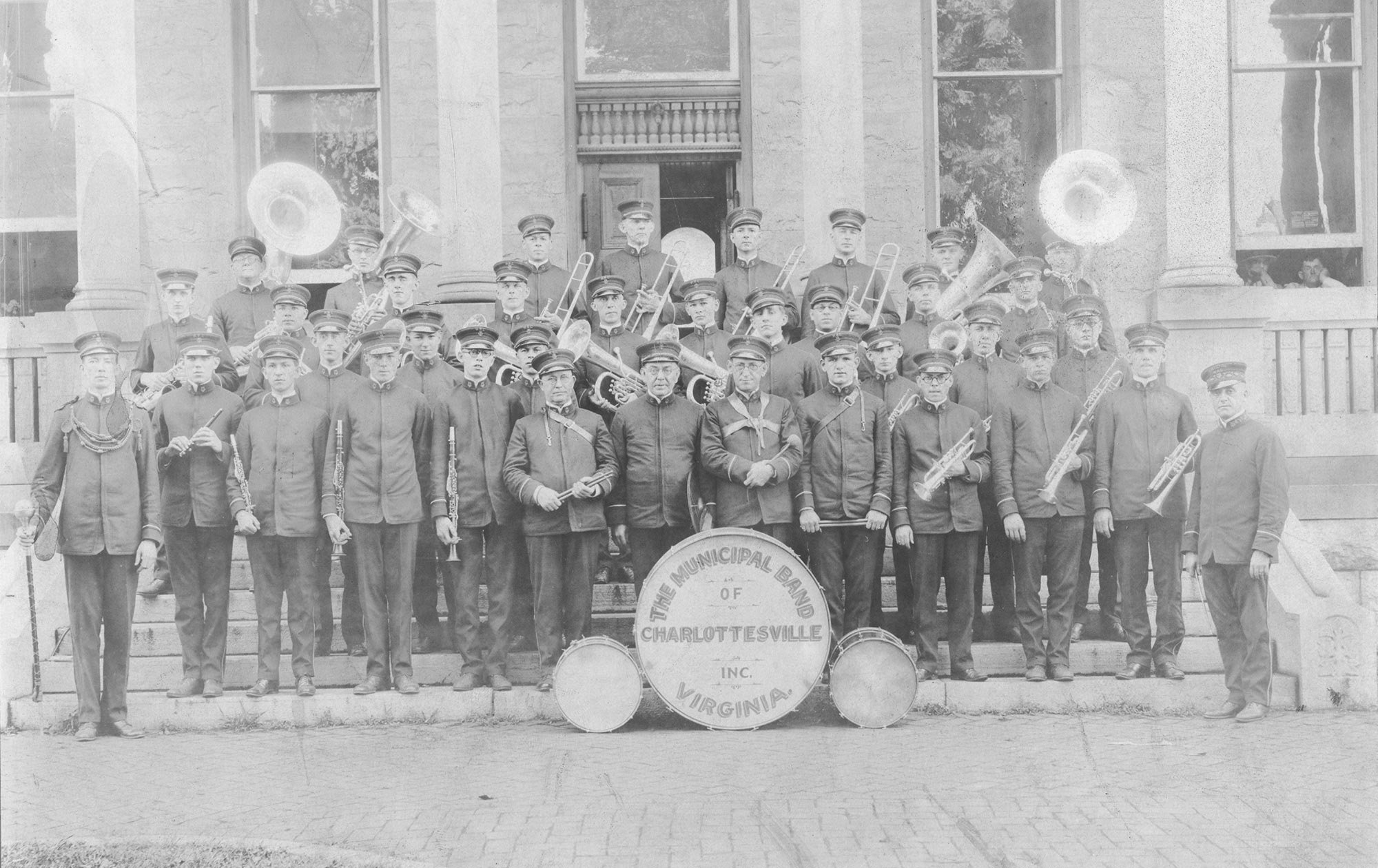 Black-and-white photo of a marching band group standing on the steps of a building, holding musical instruments like trumpets, trombones, and clarinets, with a large drum in front marked "The Municipal Band of Charlottesville, Virginia."