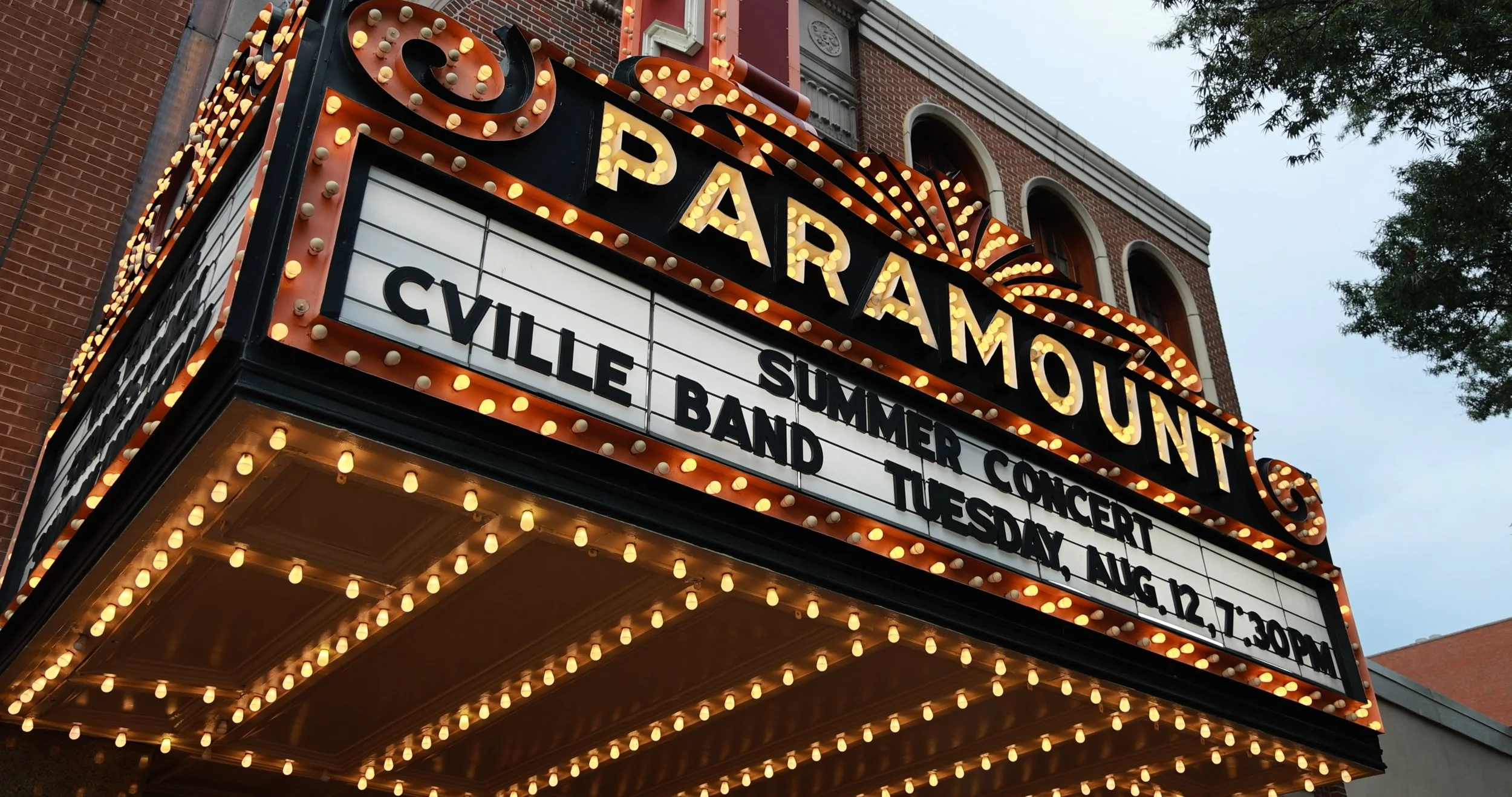 Marquam Theater marquee with bright light bulbs, displaying showtimes and events, and a brick building with arched windows in the background.