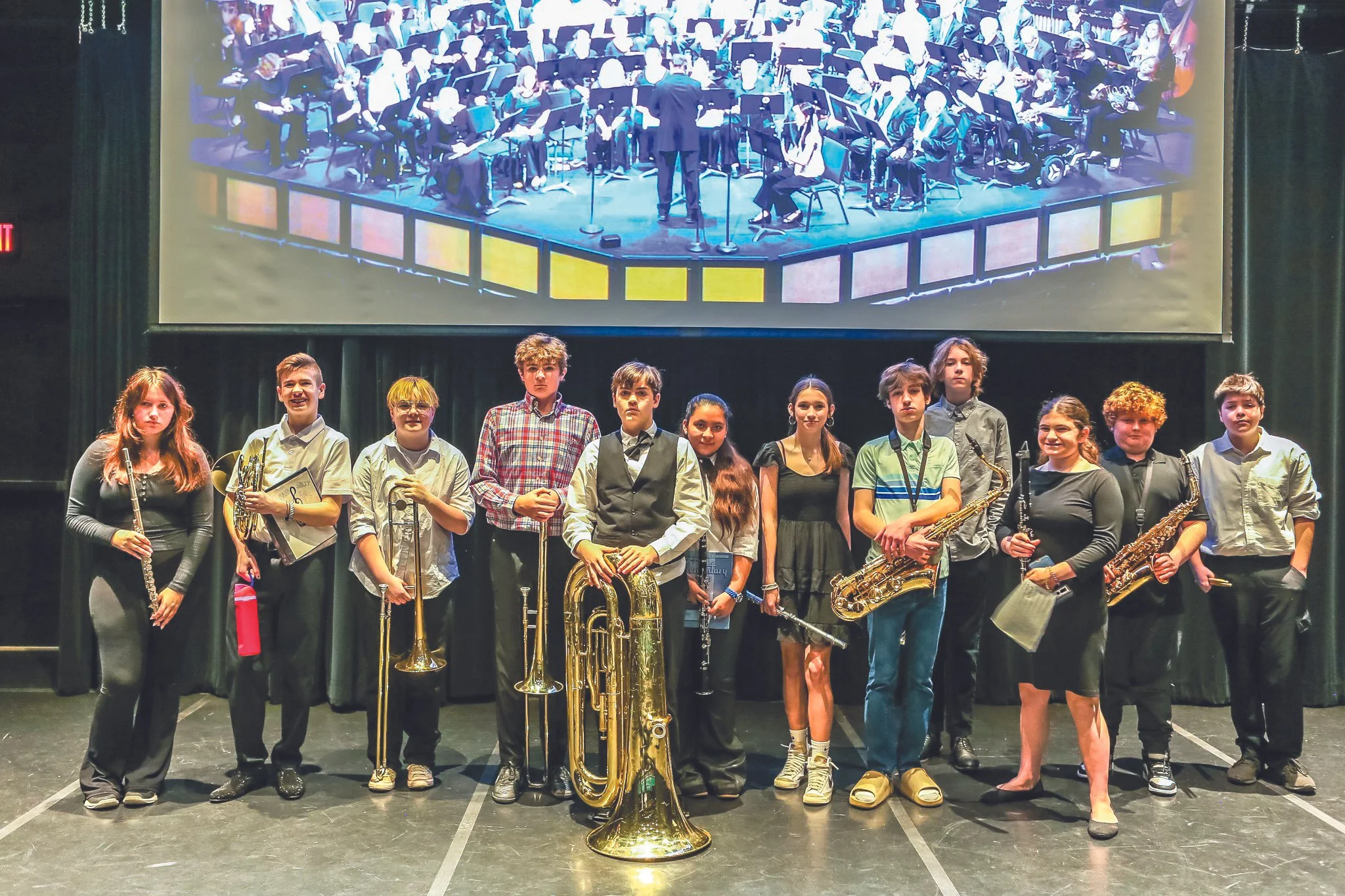 A group of teenagers holding musical instruments, standing on stage in front of a large screen displaying an orchestra. The teenagers appear to be part of a youth band or orchestra performance.