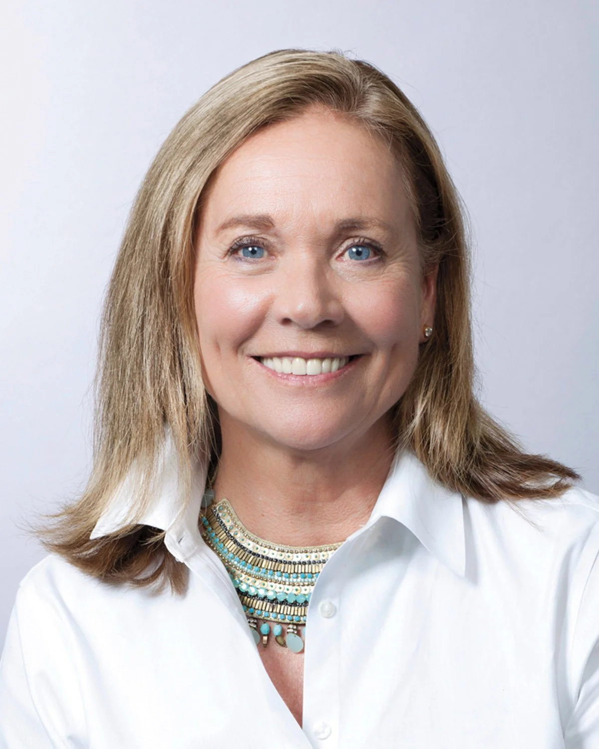A woman with shoulder-length blonde hair, blue eyes, and wearing a white collared shirt, smiling against a light gray background.