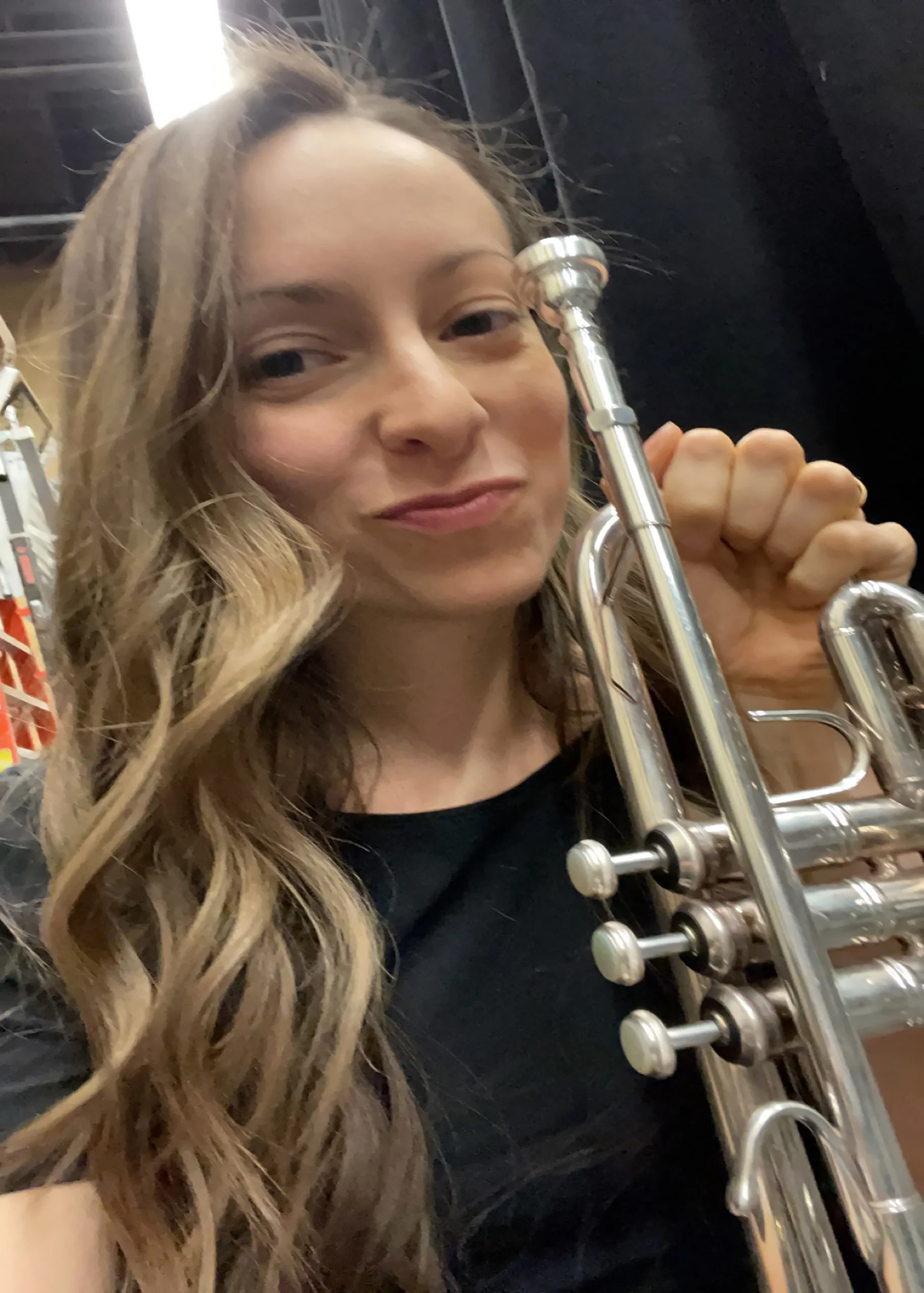 A woman with long, wavy blonde hair holding a silver trumpet and smiling indoors.